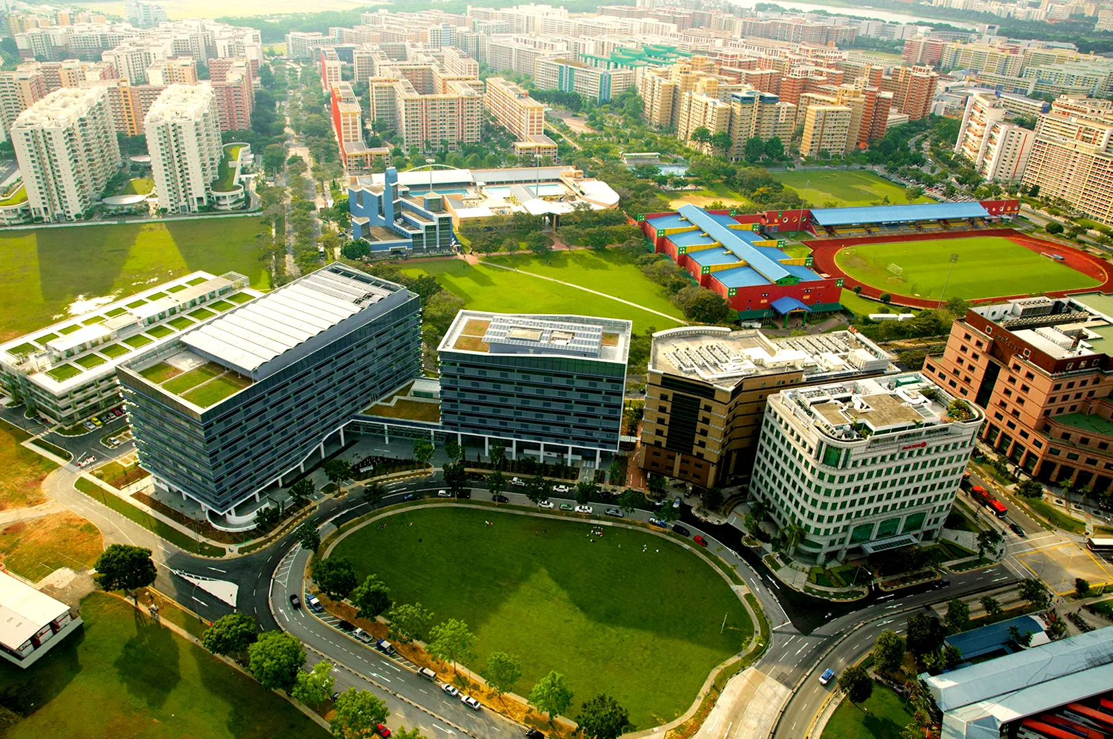 Aerial view of a modern urban campus with several multi-story buildings surrounded by green fields and a red athletic track, under a clear sky.