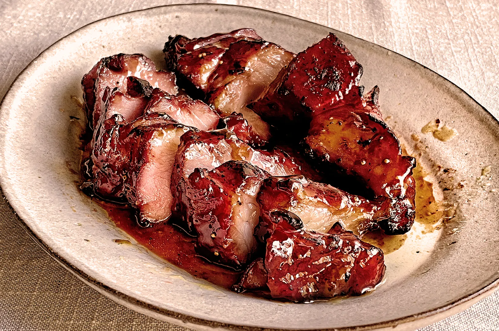 Slices of glazed barbecued pork with charred edges are arranged on a rustic ceramic plate. The pork's glossy surface suggests a savory marinade.
