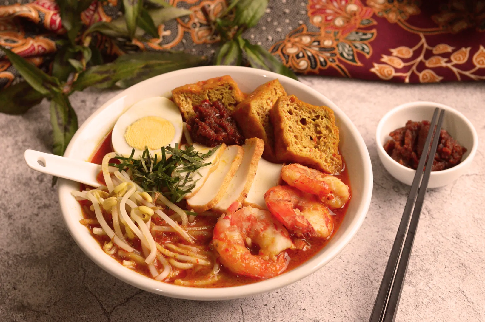 A vibrant bowl of prawn noodle soup topped with bean sprouts, herbs, half a boiled egg, and fried tofu, served with chili paste. Elegant fabric backdrop.