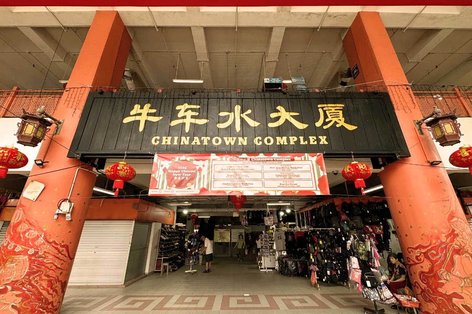 Entrance of Chinatown Complex with large black and gold sign in English and Chinese. Red lanterns hang from orange pillars. A few people walk below.