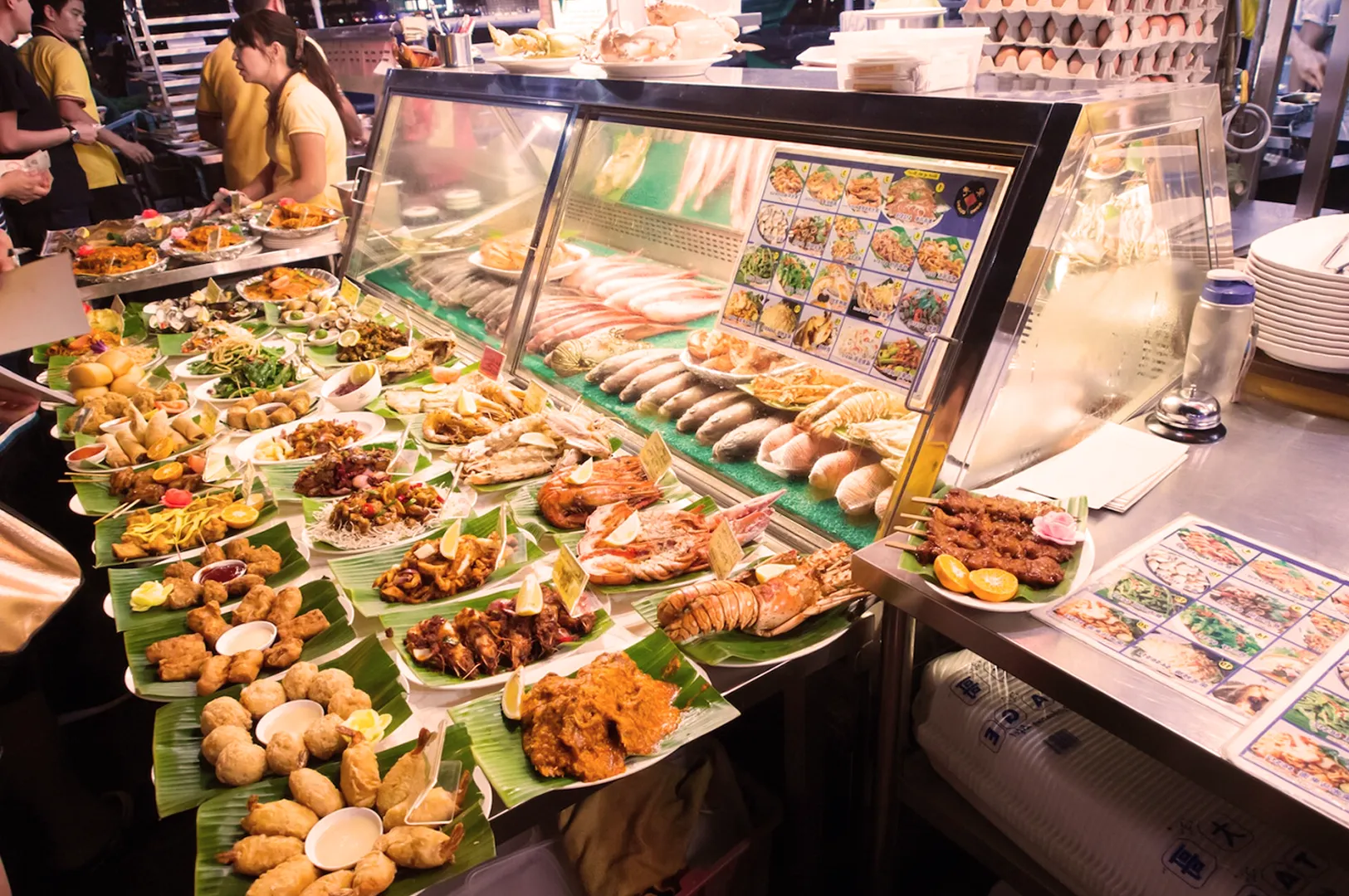 A vibrant street food stall displays a variety of colorful dishes including seafood, skewers, and appetizers on banana leaves, exuding a lively, bustling atmosphere.