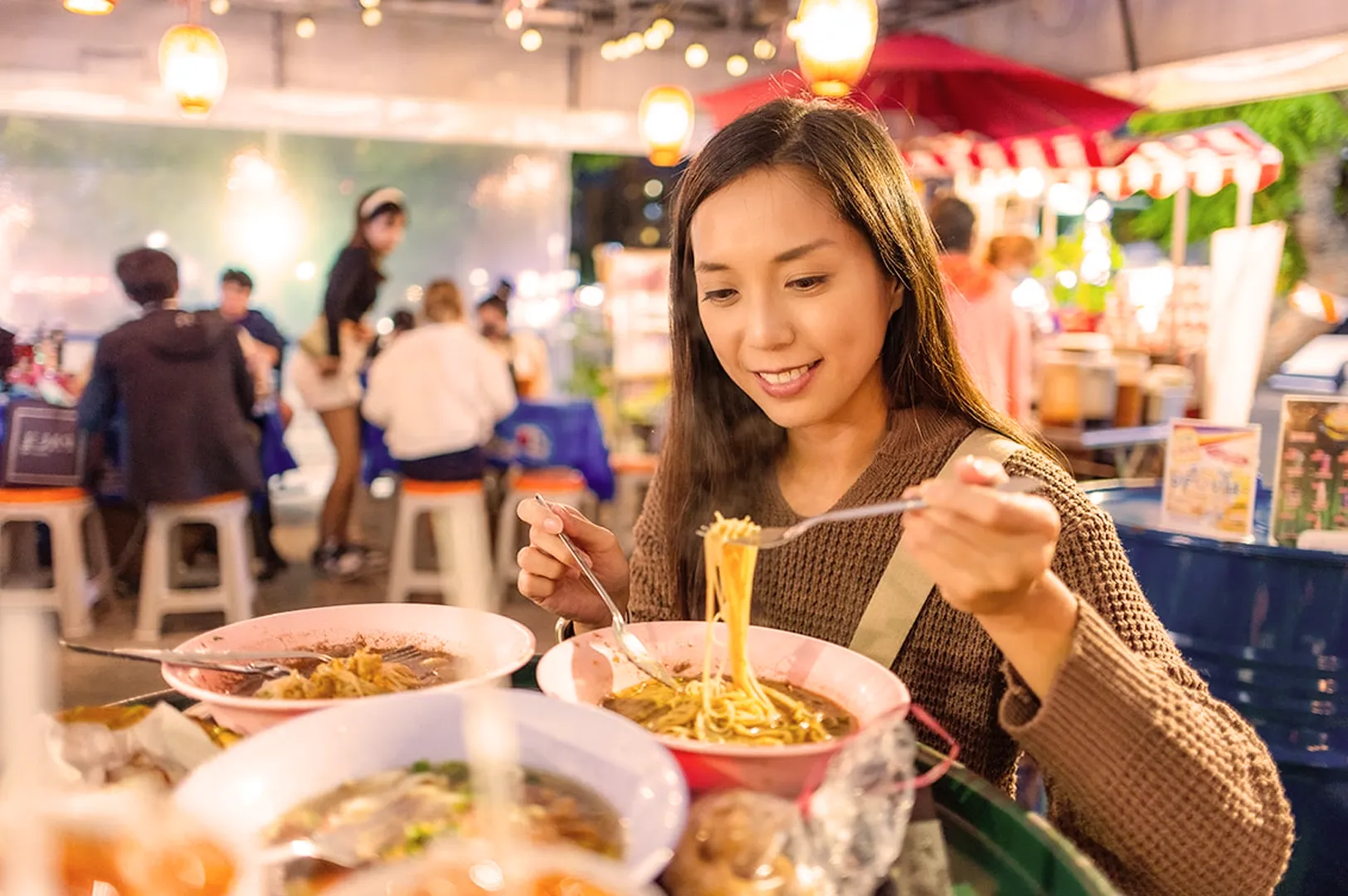 A woman happily eating noodles at a vibrant, outdoor food market. She is surrounded by colorful bowls of soup, with warm lighting and people in the background.