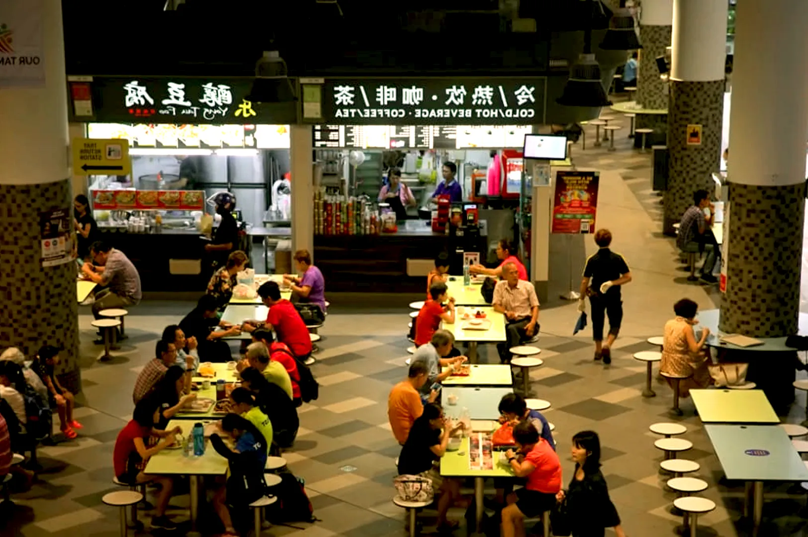 A bustling hawker center with people sitting at shared tables, eating and chatting. Bright signage reads "Cold, Hot Beverage." The atmosphere is lively.