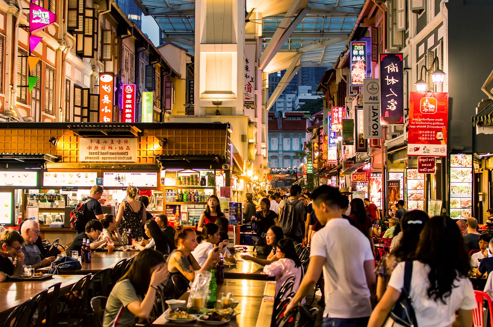 Bustling night market with people dining outdoors along a vibrant street lined with illuminated signs and food stalls, creating a lively atmosphere.