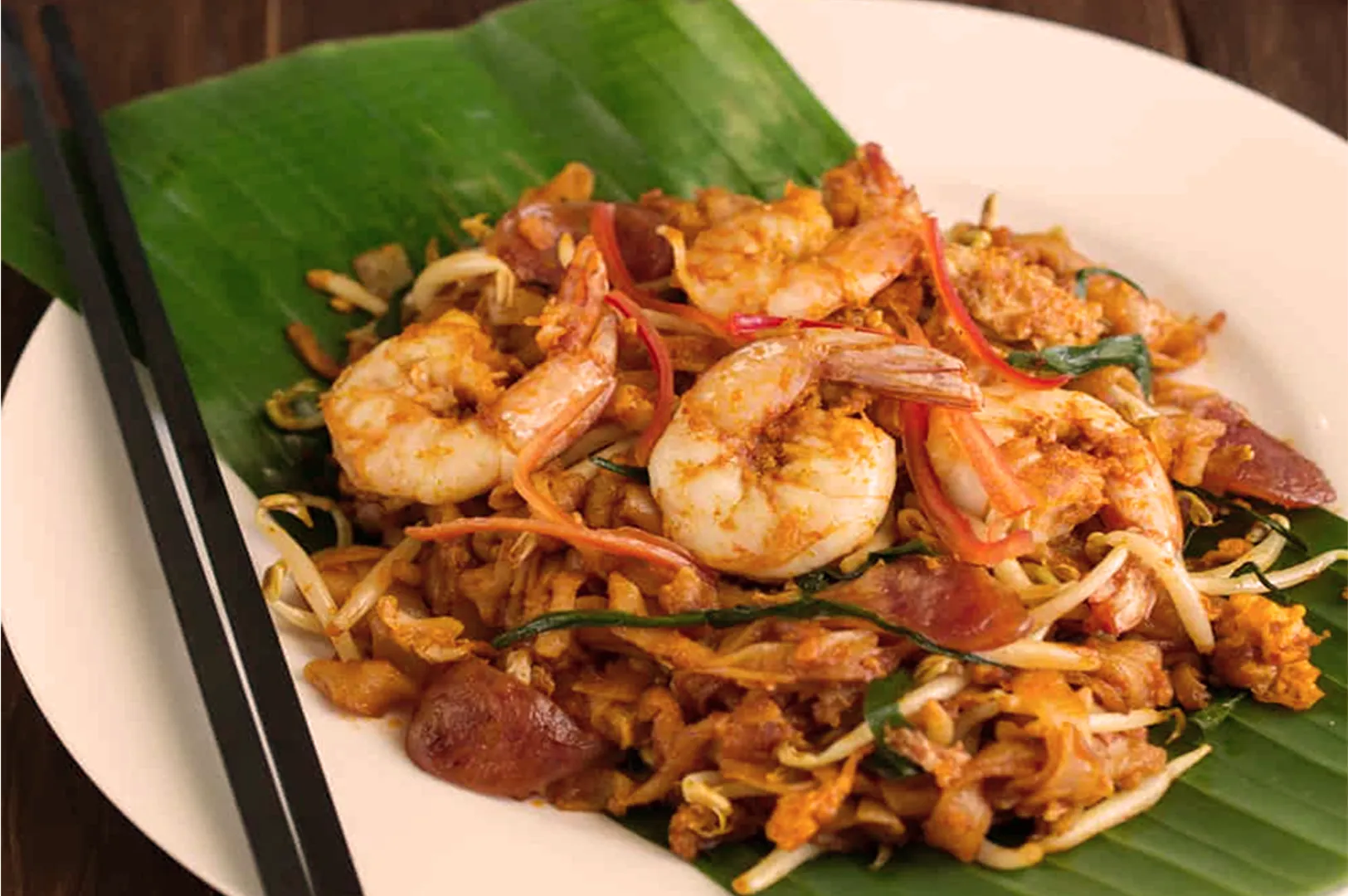 A vibrant plate of Pad Thai featuring shrimp, bean sprouts, and noodles on a green banana leaf. Black chopsticks are placed beside, evoking an inviting, aromatic appeal.