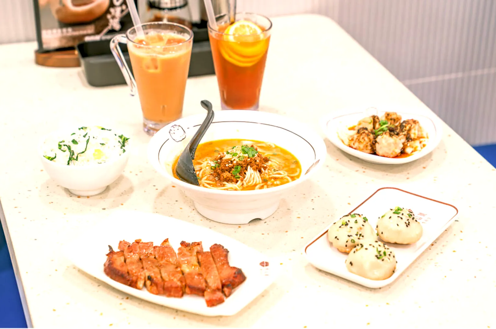 A brightly lit table with a bowl of noodle soup at the center, surrounded by meats, dumplings, and rice dishes. Iced teas are placed in the background.