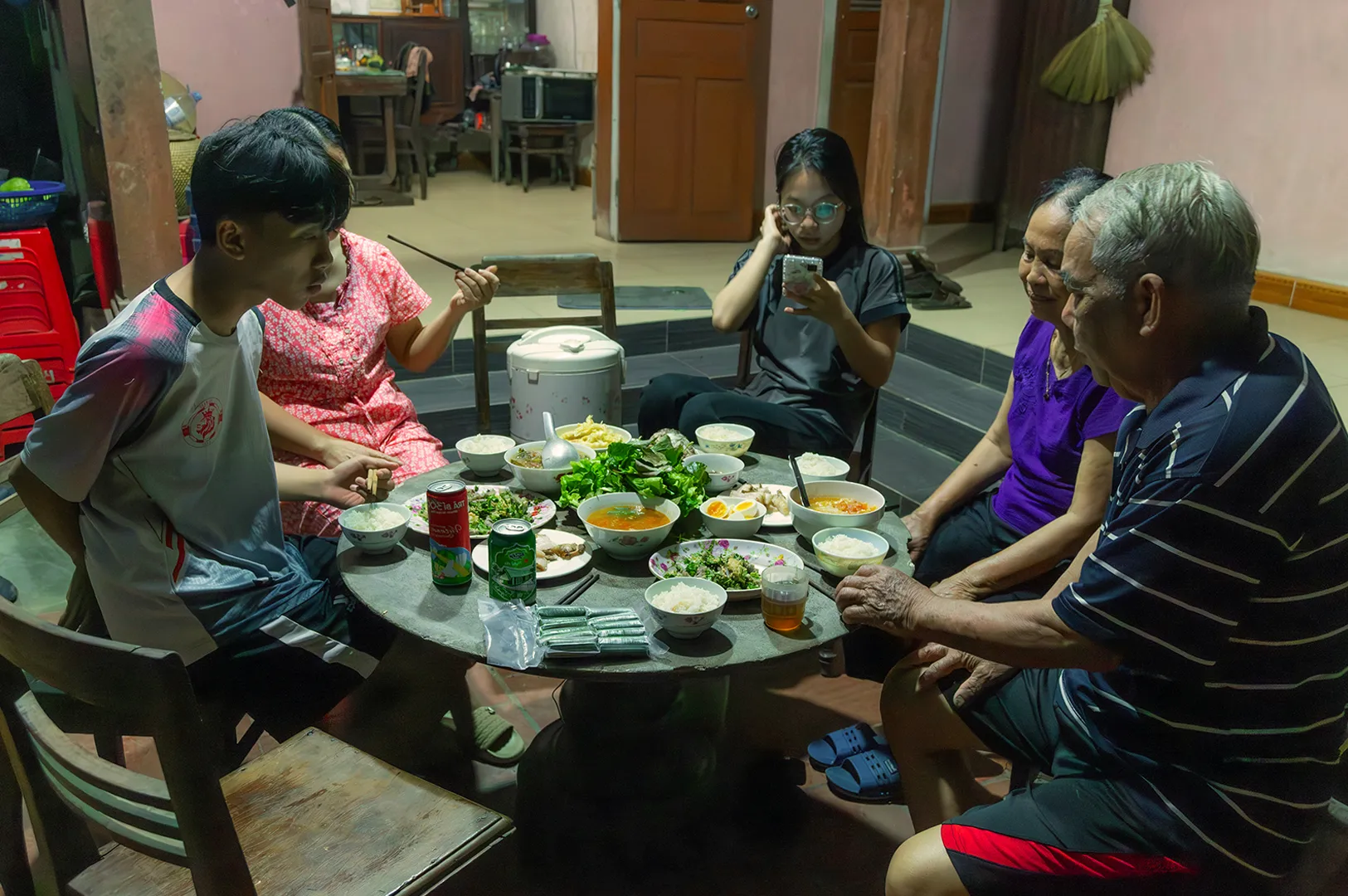 A family of five gathers around a small table filled with various dishes, rice, and drinks in a warmly lit room. The atmosphere is casual and intimate.