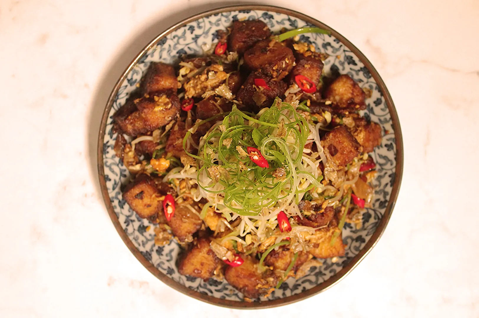 A bowl of stir-fried radish cake topped with green onions, bean sprouts, and red chili slices on a patterned blue and white plate, creating a vibrant and appetizing presentation.