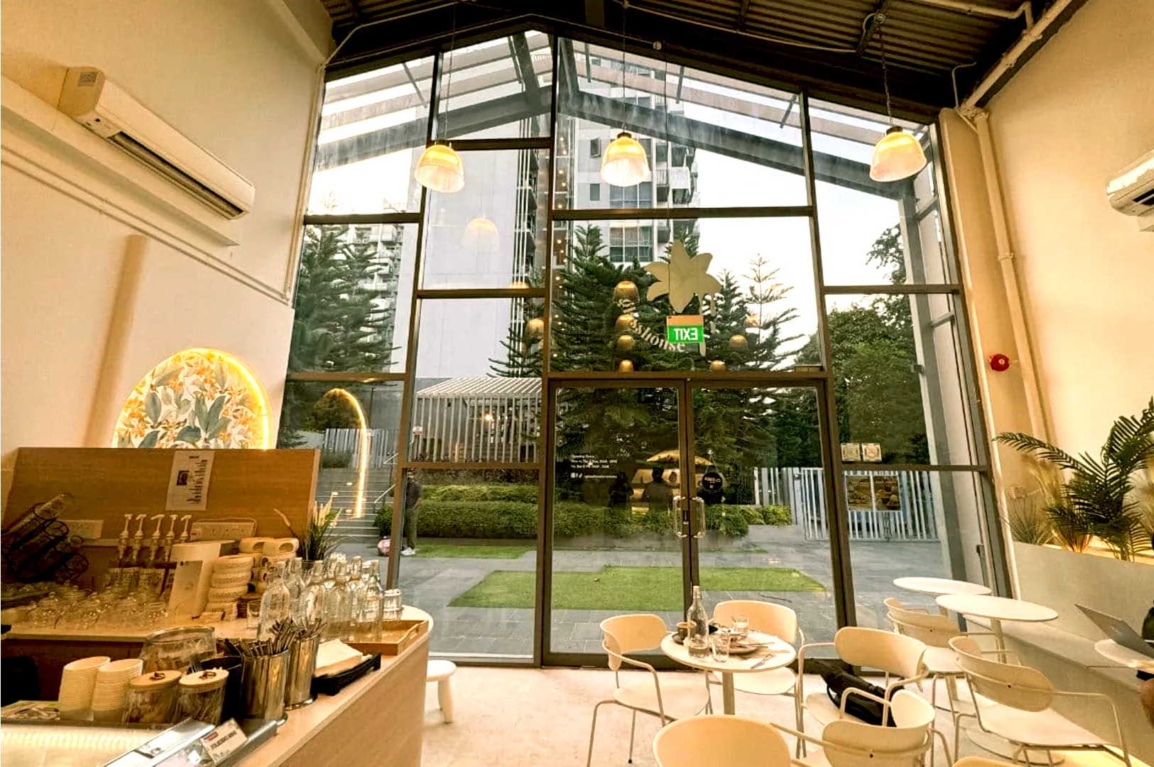 Chic cafe with high glass walls, white chairs, and tables. Warm lighting creates a cozy ambiance. Outside, greenery and tall buildings are visible.
