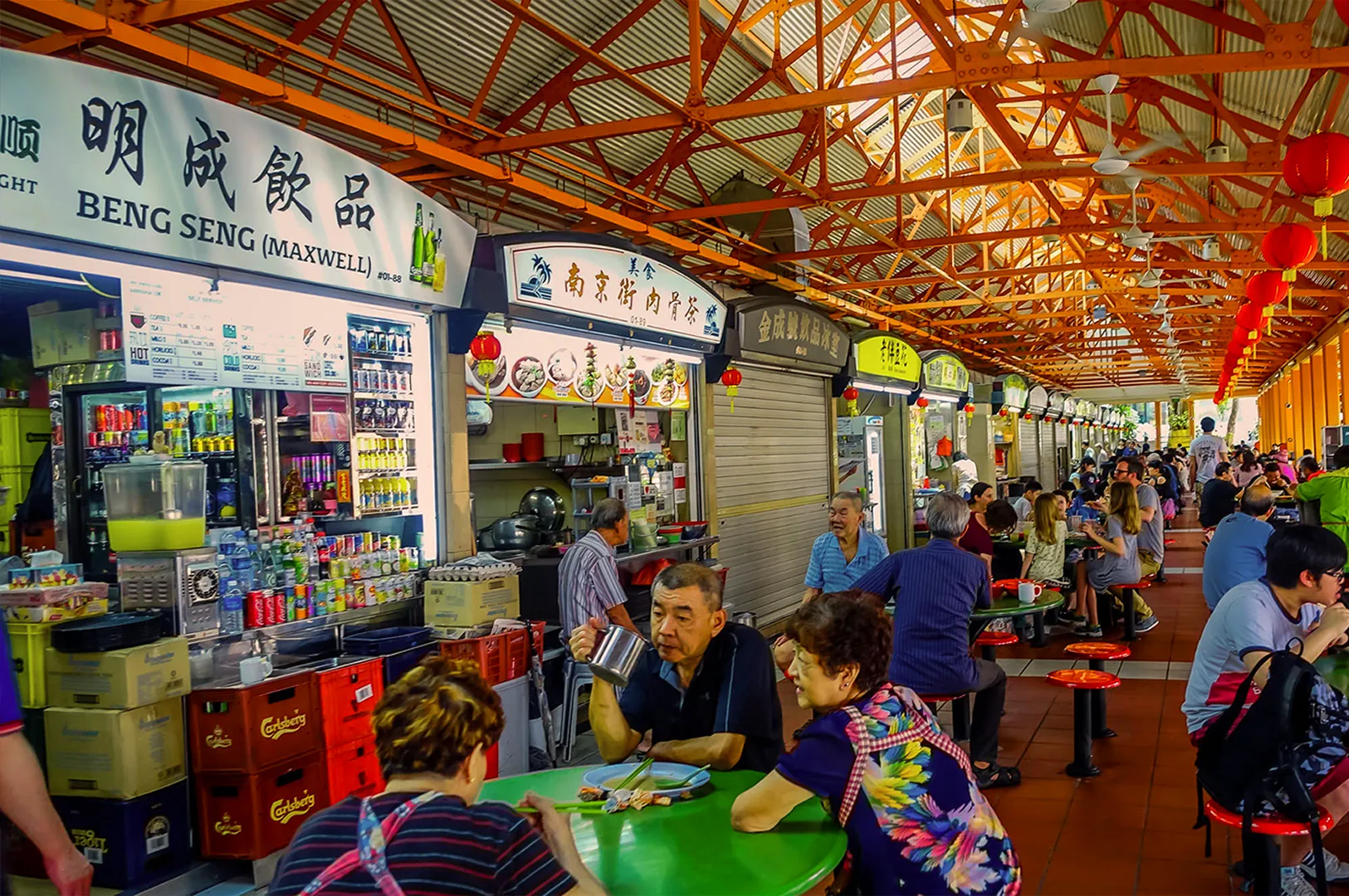 Bustling hawker center with people dining at colorful tables under an orange lattice roof. Food stalls line the background, creating a lively atmosphere.