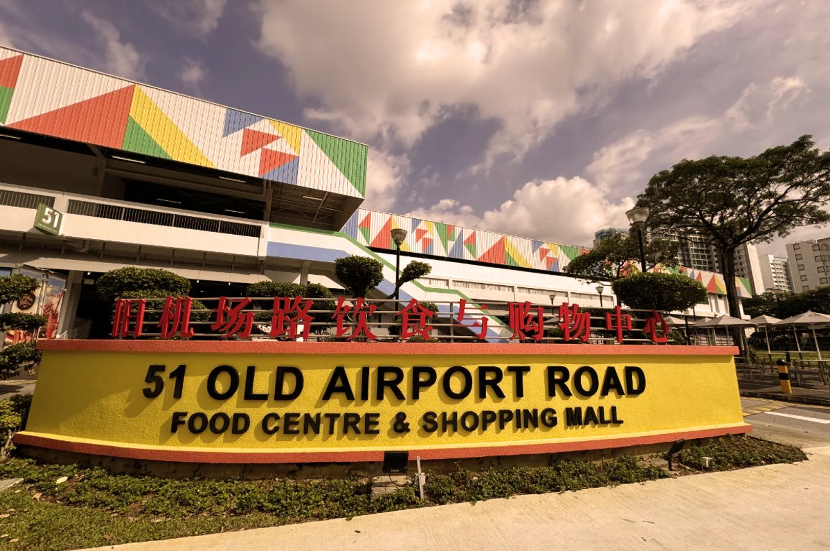 Sign for 51 Old Airport Road Food Centre & Shopping Mall under a colorful building facade with geometric patterns. Vibrant and lively atmosphere.