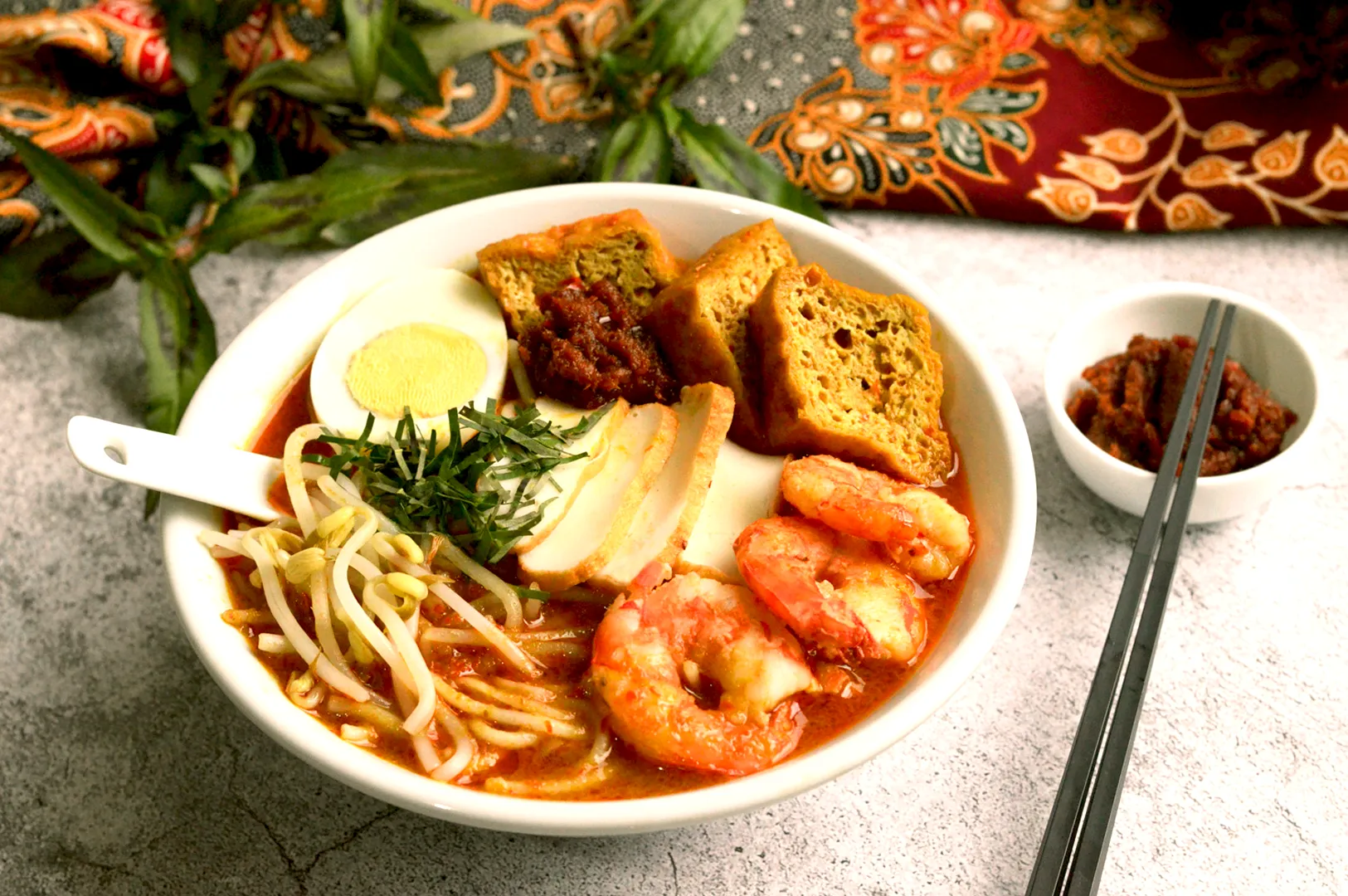 A vibrant bowl of prawn noodle soup topped with bean sprouts, herbs, half a boiled egg, and fried tofu, served with chili paste. Elegant fabric backdrop Laksa
