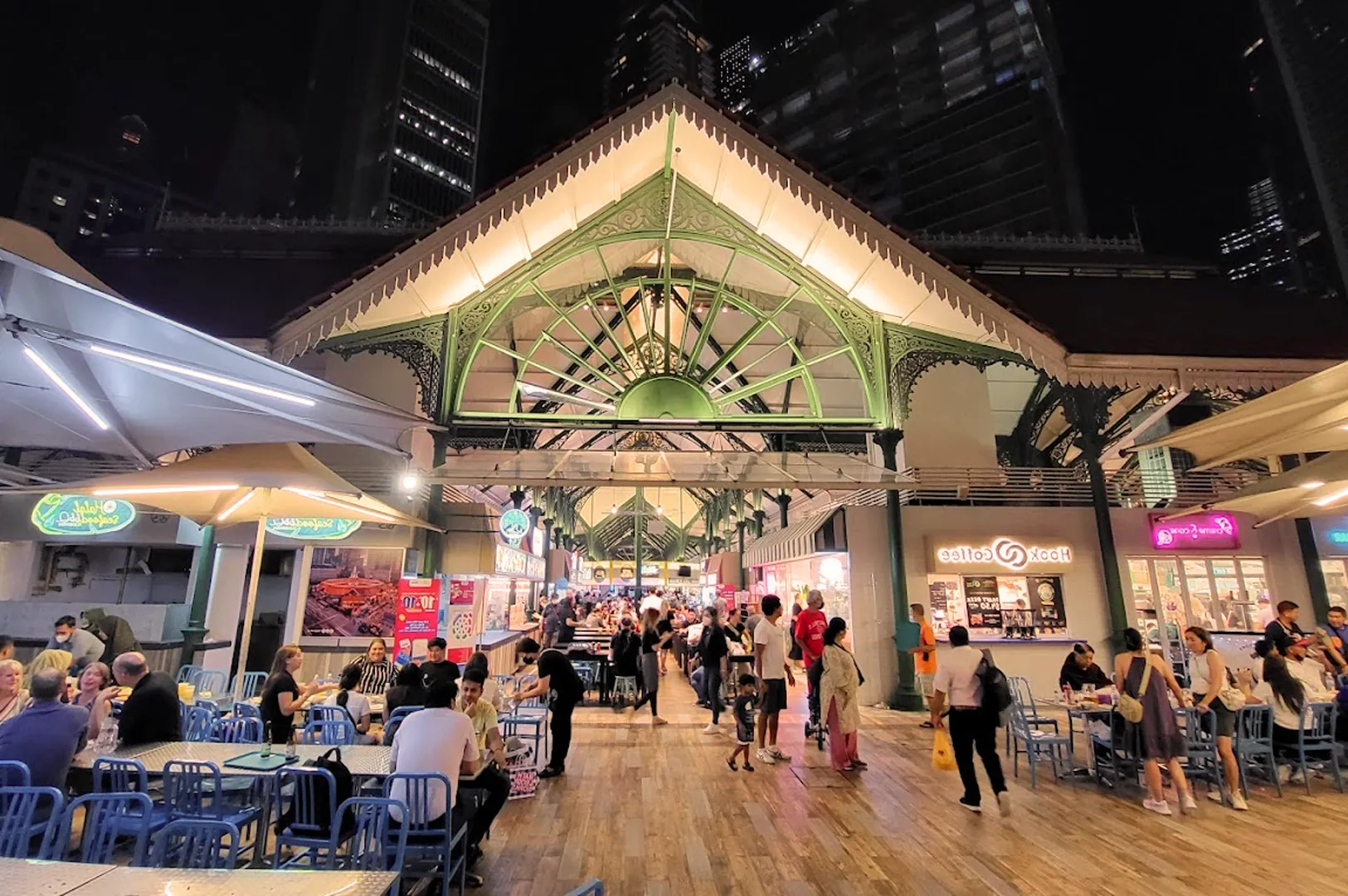 Bustling night food market with a peaked, green architectural structure. Crowds sit at blue tables, enjoying meals under bright lights, creating a lively, social atmosphere.