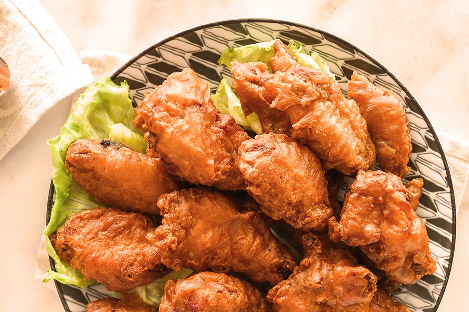 A plate of crispy, golden-brown fried chicken wings on a bed of fresh lettuce. The dish is arranged on an elegant patterned platter, creating a savory and inviting presentation.