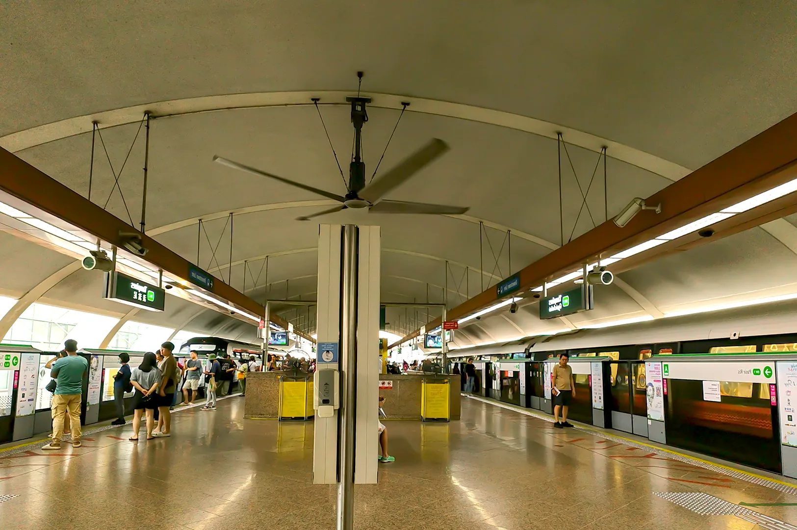 Spacious subway platform with passengers waiting, curved ceiling, central large fan, overhead signs, trains on both sides. Calm atmosphere.