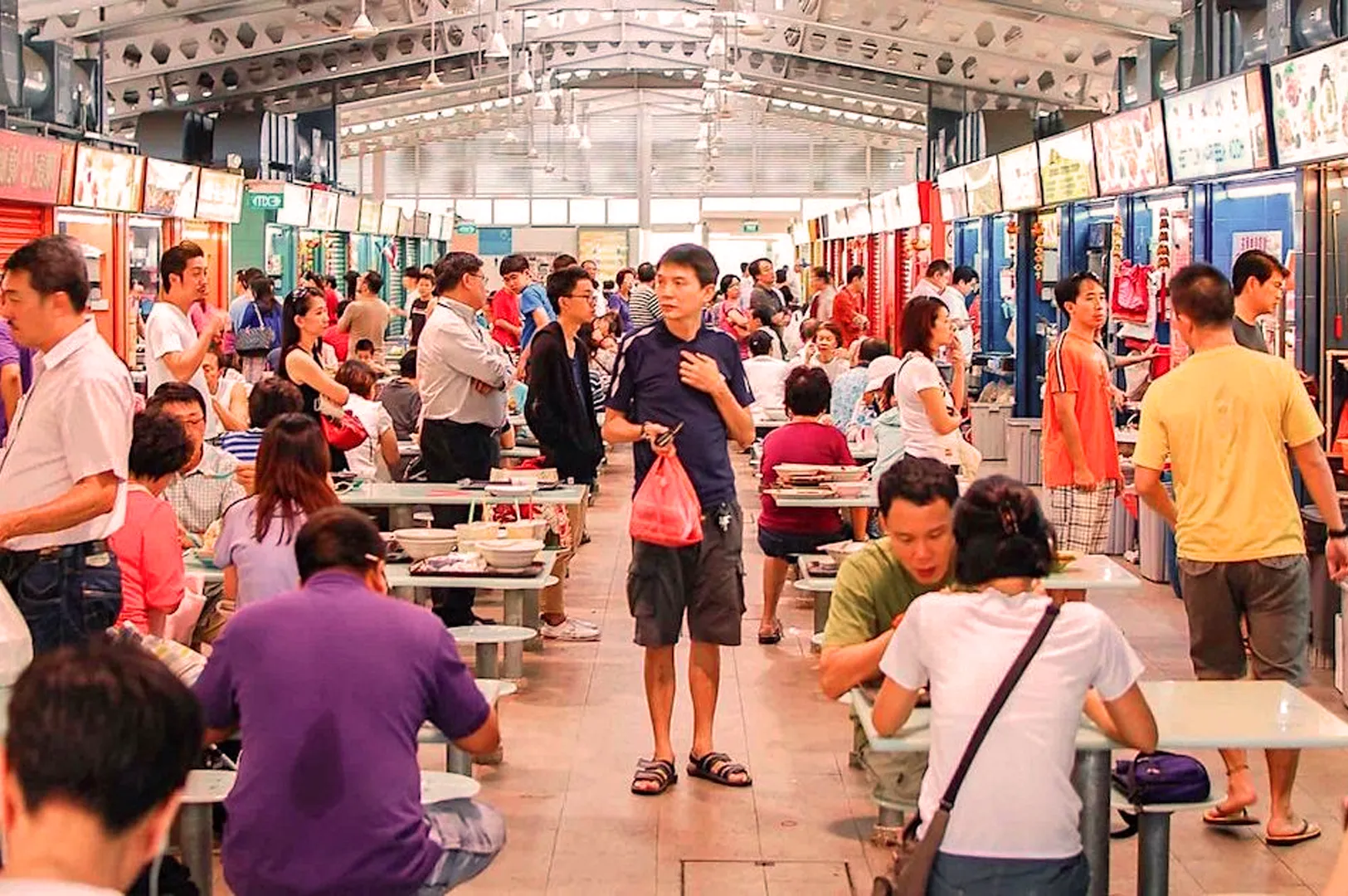 Bustling food market scene with diverse people seated at tables eating and socializing. Vendors serve food under brightly lit stalls in a lively atmosphere.