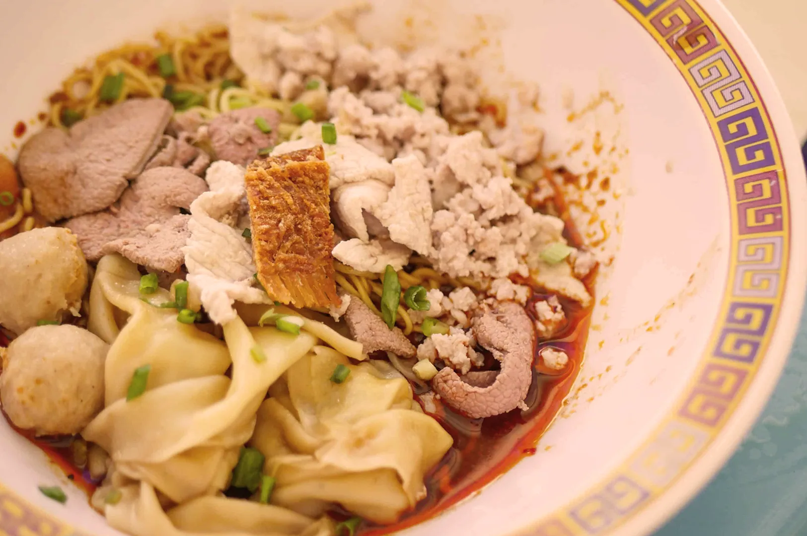A colorful bowl of Asian noodle soup with dumplings, meat slices, and fresh herbs, set against a decorative patterned rim. The dish looks flavorful and inviting.
