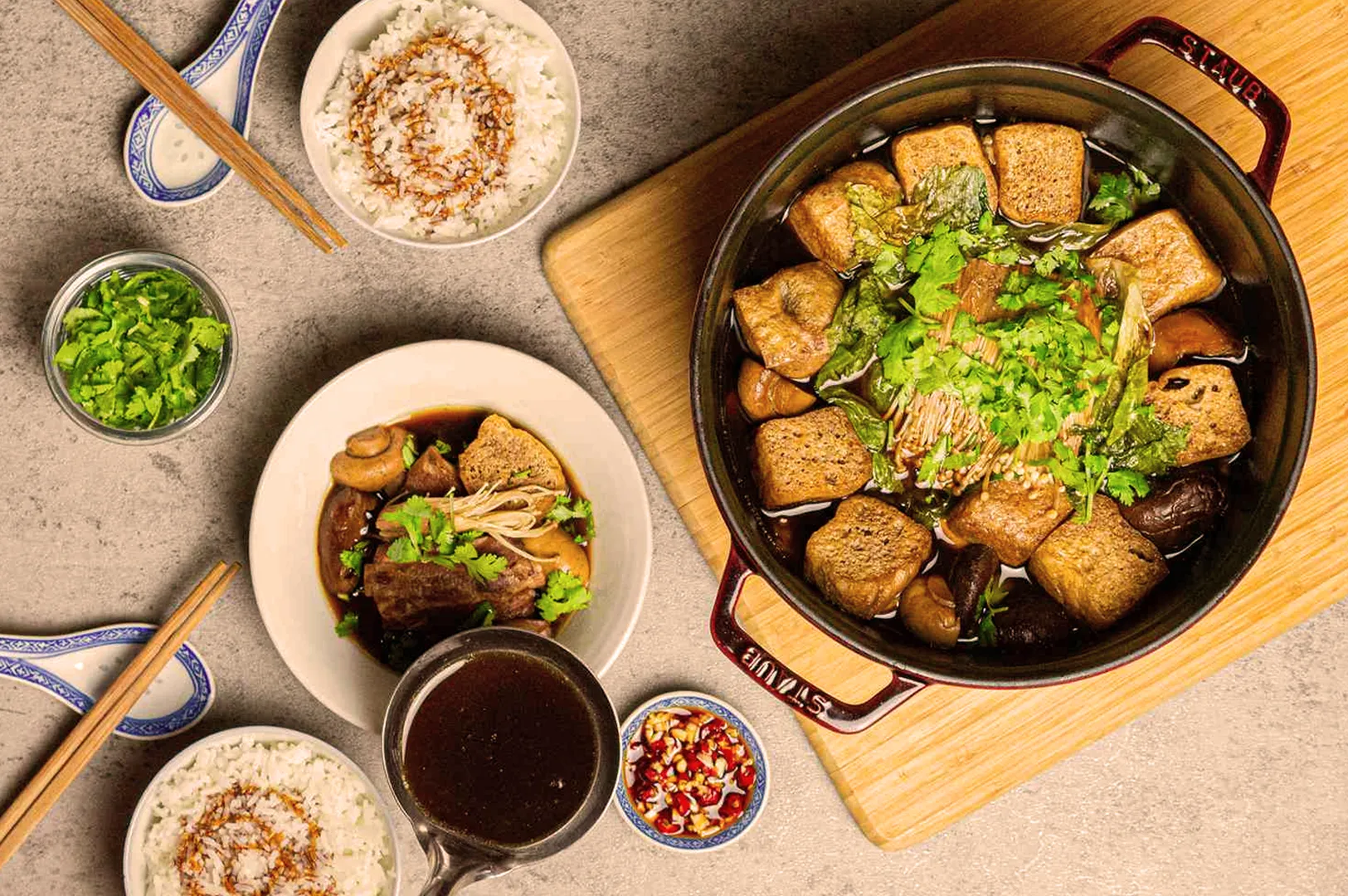 A vibrant Asian hotpot filled with tofu, mushrooms, and greens on a wooden board. Surrounding dishes include rice, sauce, and fresh herbs, conveying a cozy and communal dining atmosphere.