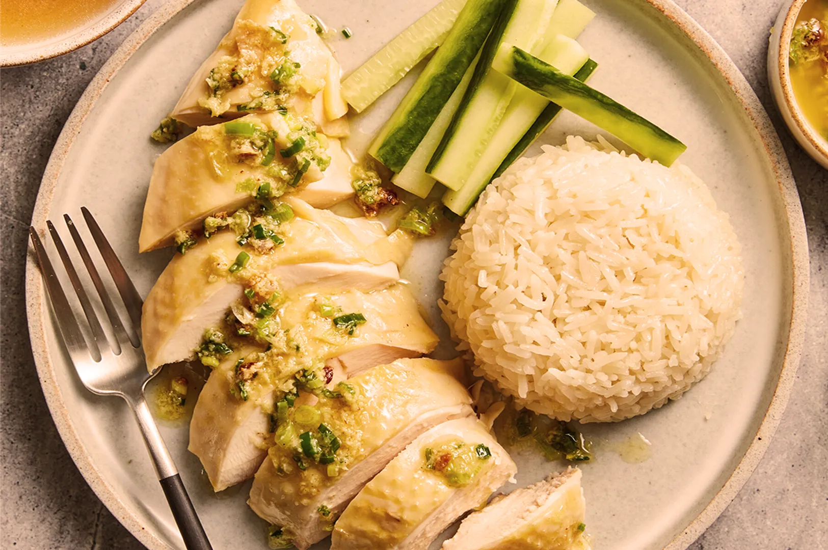 Plate of sliced poached chicken with green onion sauce, cucumber strips, and a mound of white rice. A fork lies beside the chicken.