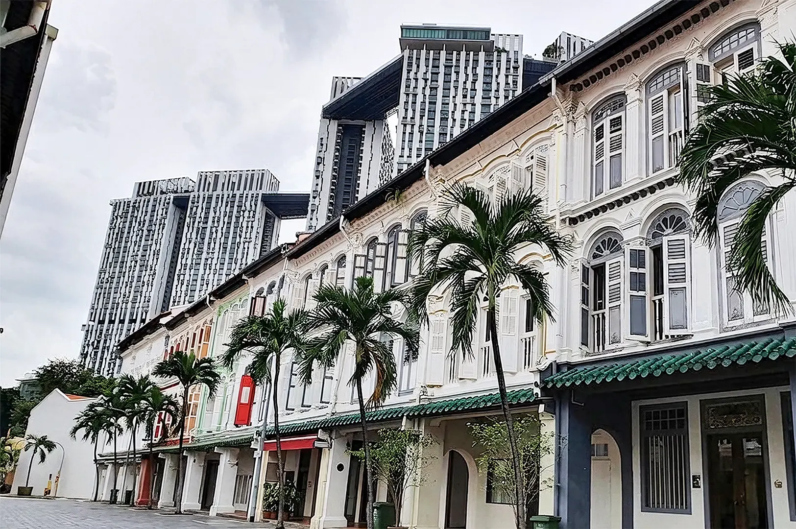 A historic white shophouses with green-tiled roofs and palm trees in Tanjong Pagar, framed against the modern, towering skyscrapers of Pinnacle@Duxton in the background.