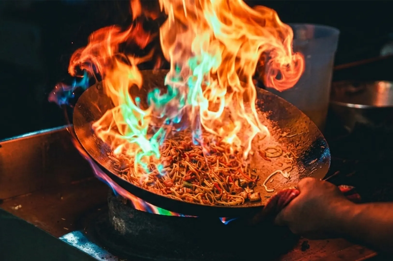 A close-up of 'wok hei' action, with noodles being tossed through soaring orange and turquoise fire.