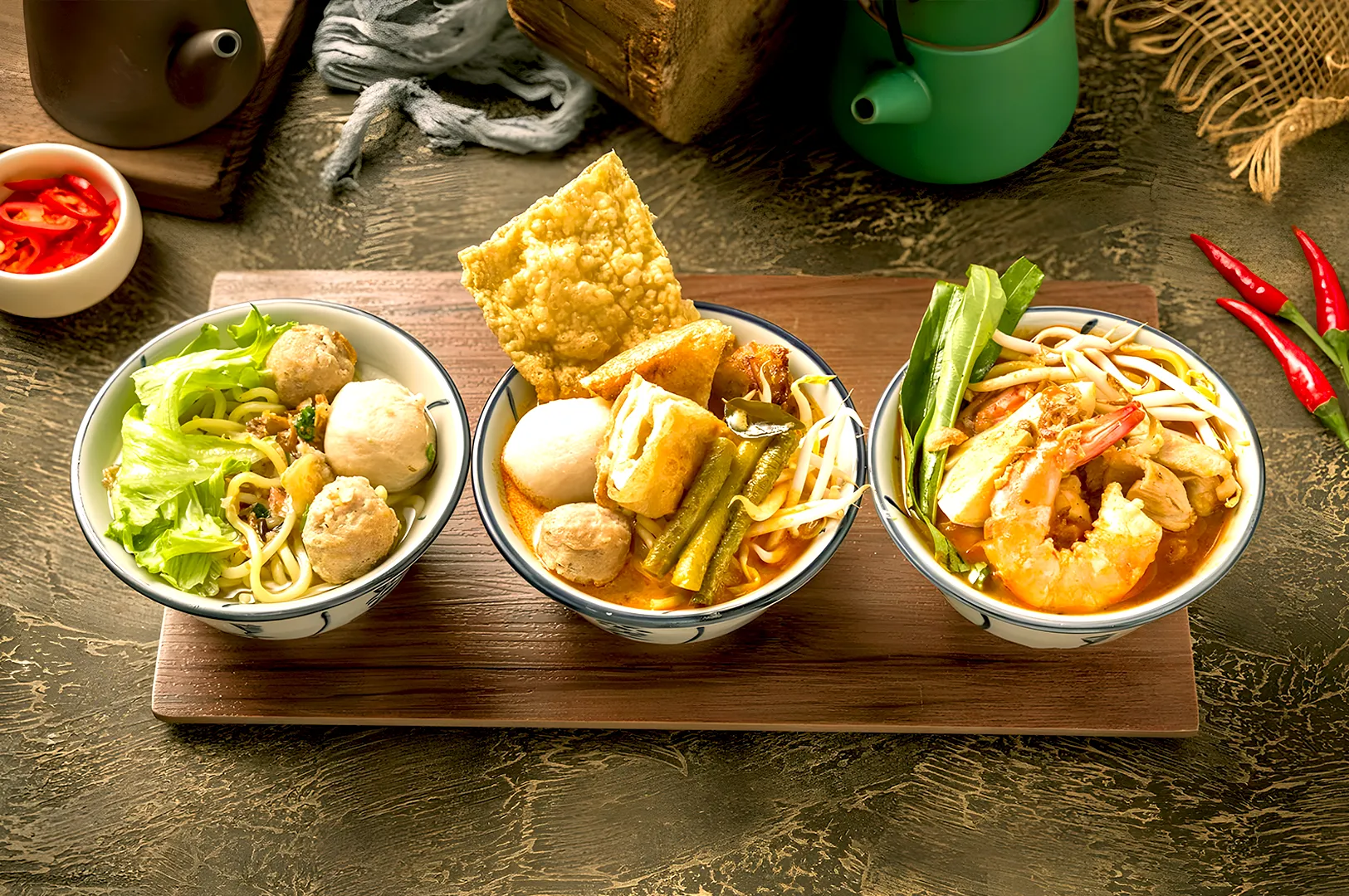 Three bowls of Chinese noodles on a wooden board. Each bowl has unique toppings, including greens, shrimp, and meatballs, with vibrant colors.