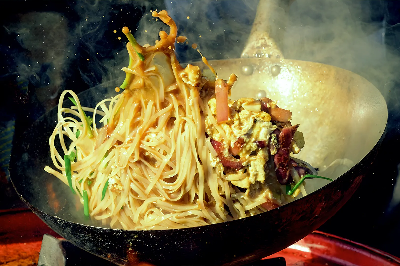 A dynamic image of Pad Thai being cooked in a wok. Noodles are tossed with splashing sauce, vegetables, and tofu, surrounded by steam.