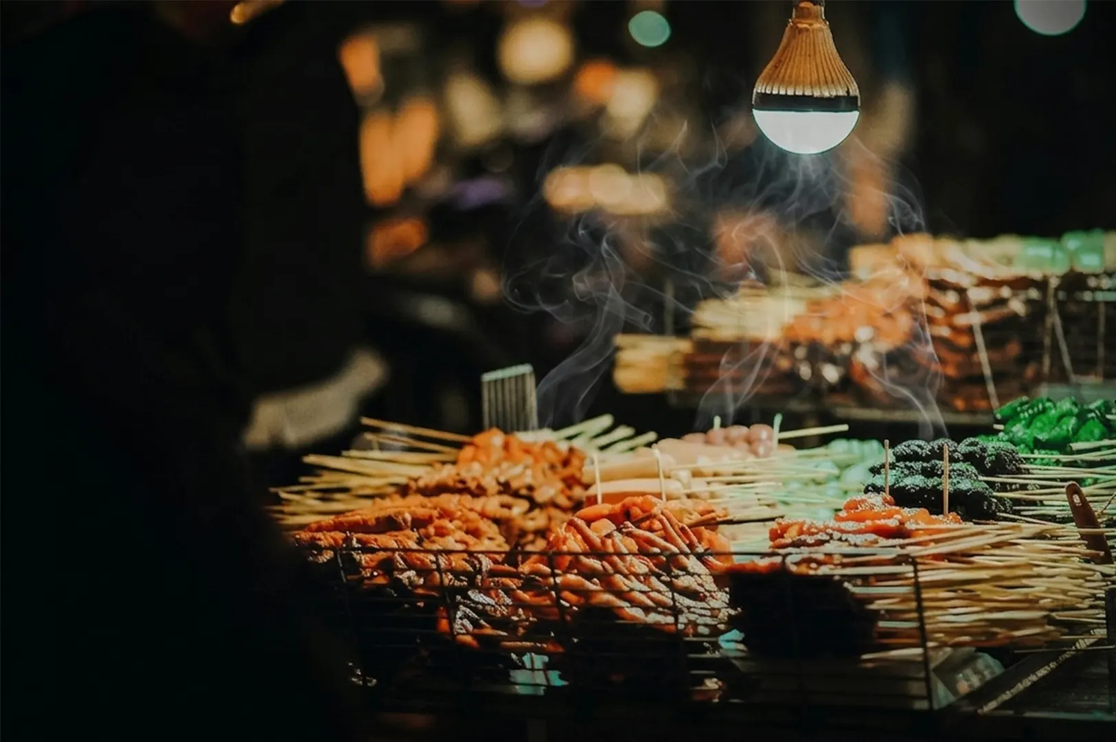 Assorted street food skewers in a wire basket with smoke under a warm hanging light at night.