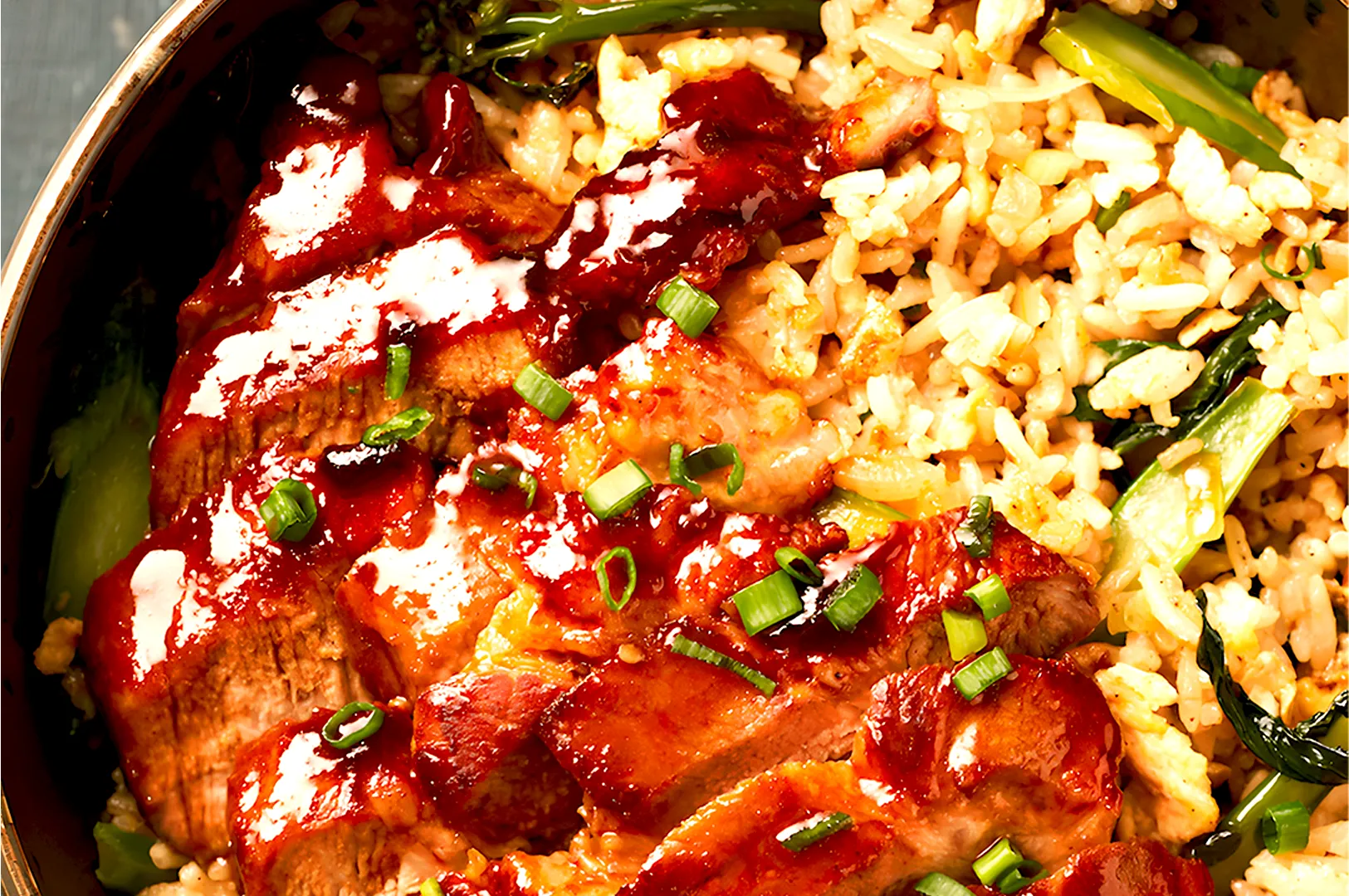 Grilled pork slices topped with sauce and green onions on fried rice with leafy greens in a bowl, conveying a savory and hearty meal.