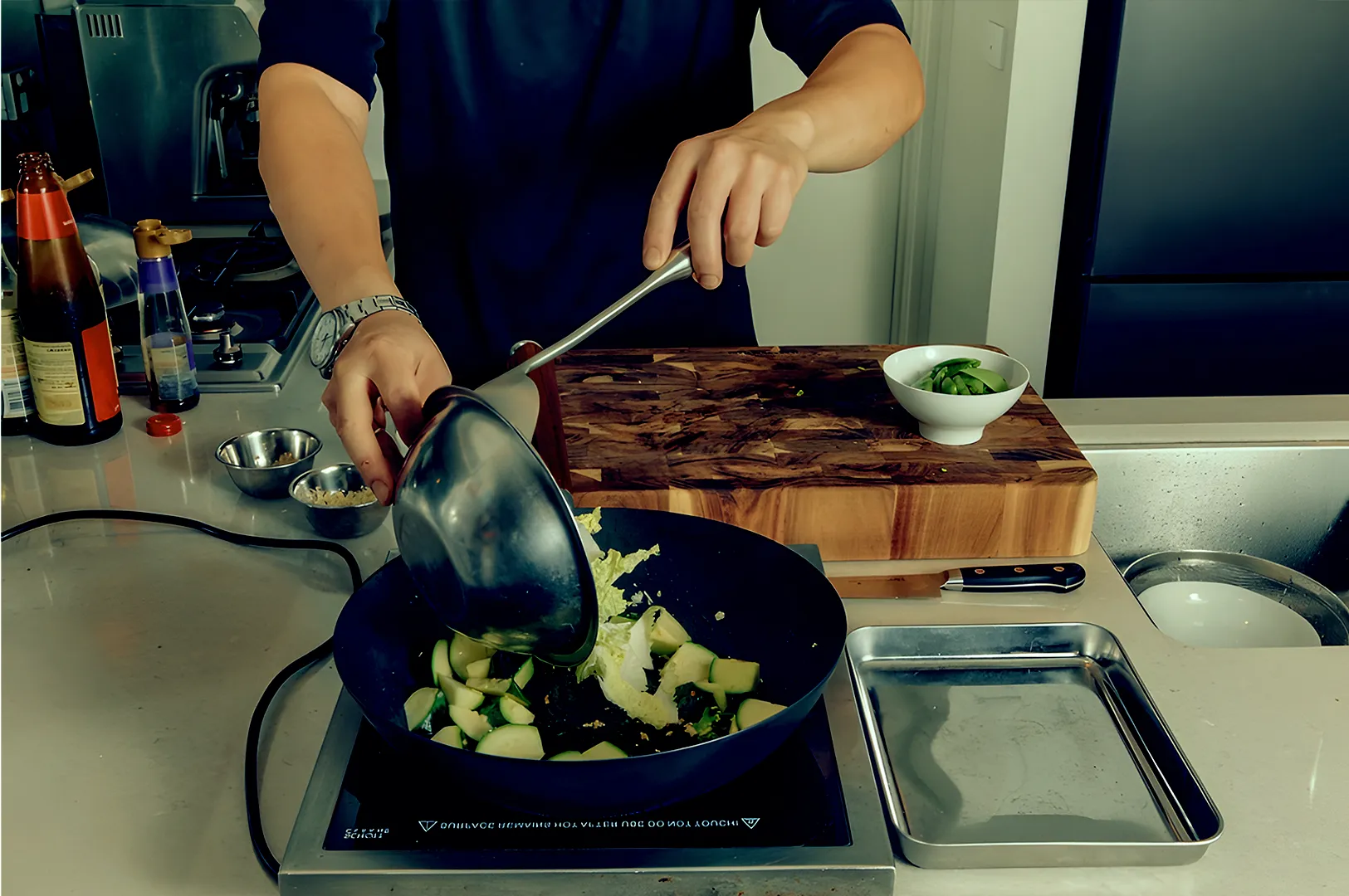 Person cooking zucchini in a black pan, pouring ingredients from a metal bowl. Countertop with cutting board, small bowl, and condiments nearby.