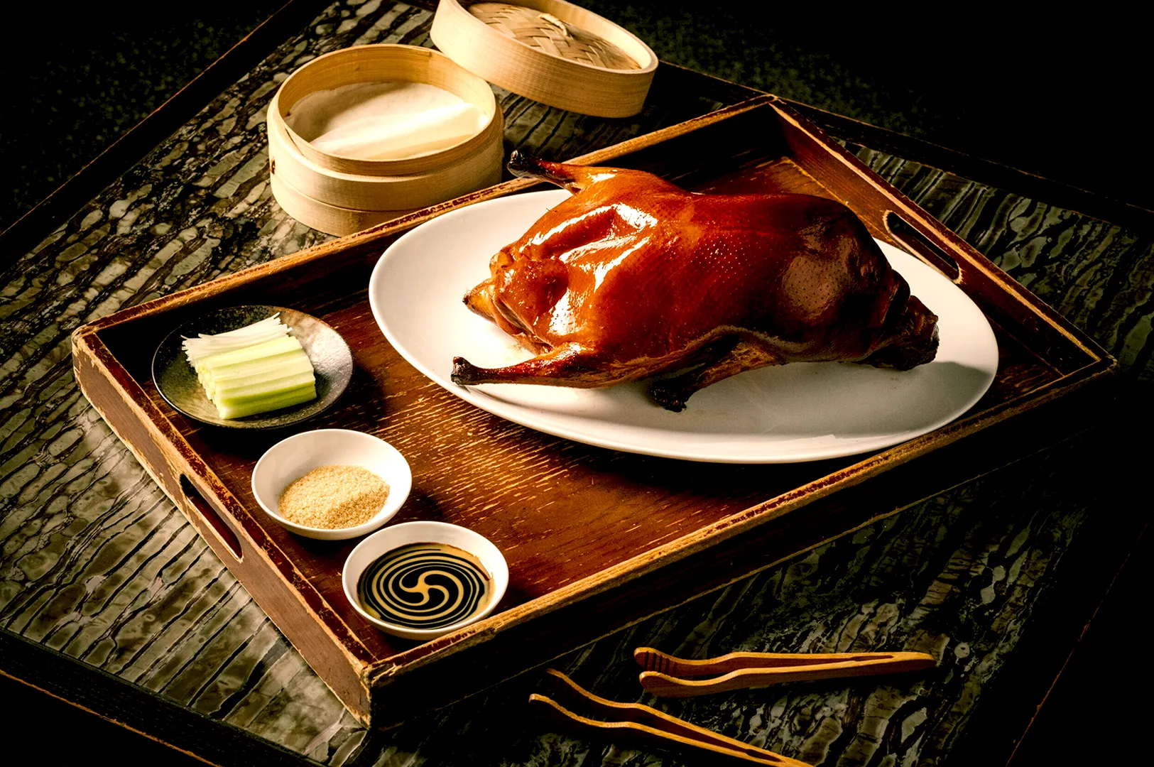 Succulent roasted duck on a white plate, surrounded by cucumber slices, sauces, and pancakes in a wooden tray. Elegant, gourmet dining setup.