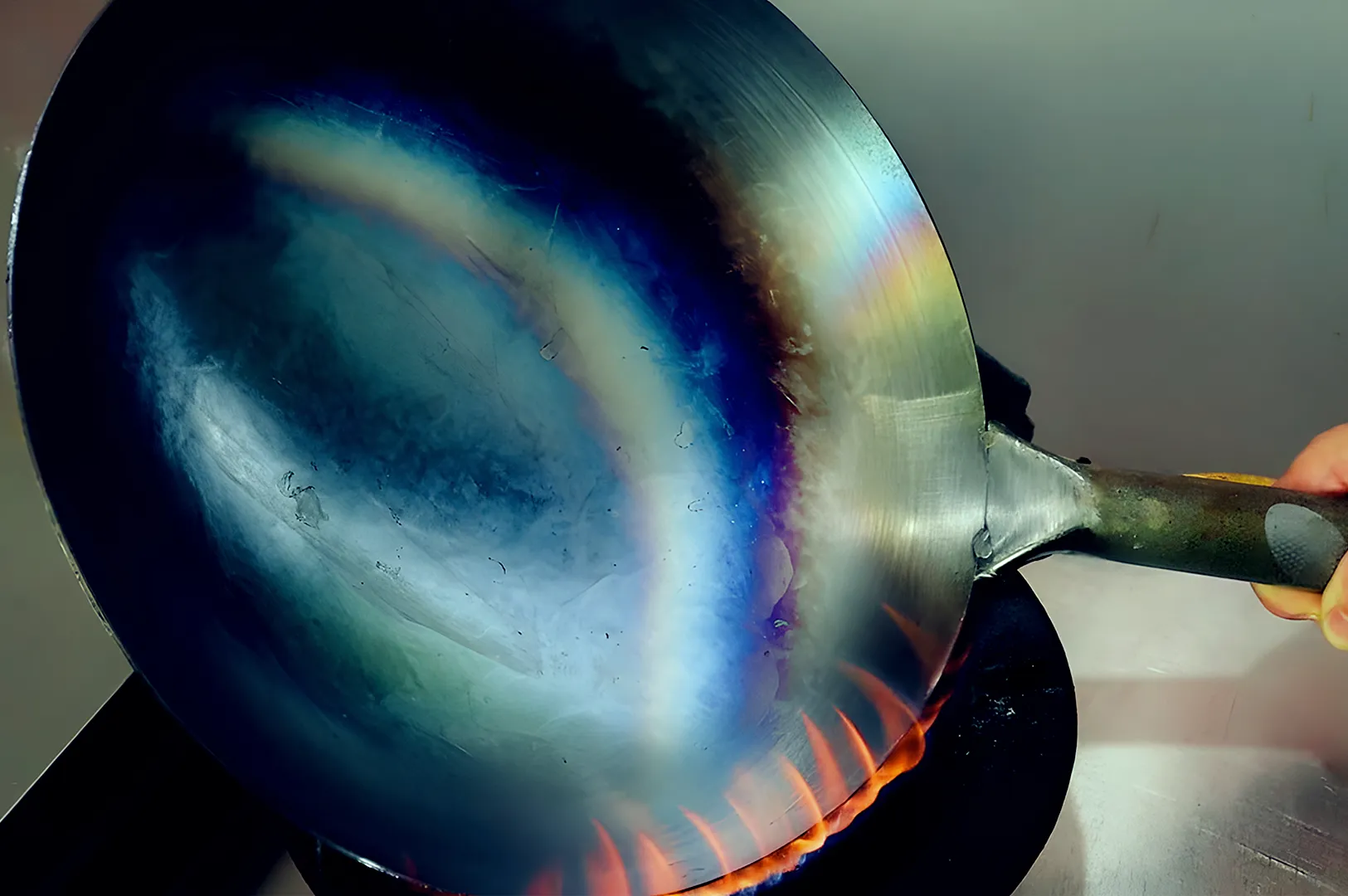 A metal wok with a rainbow patina is held over a lit gas stove. Blue, purple, and orange hues are visible, suggesting heat treatment.