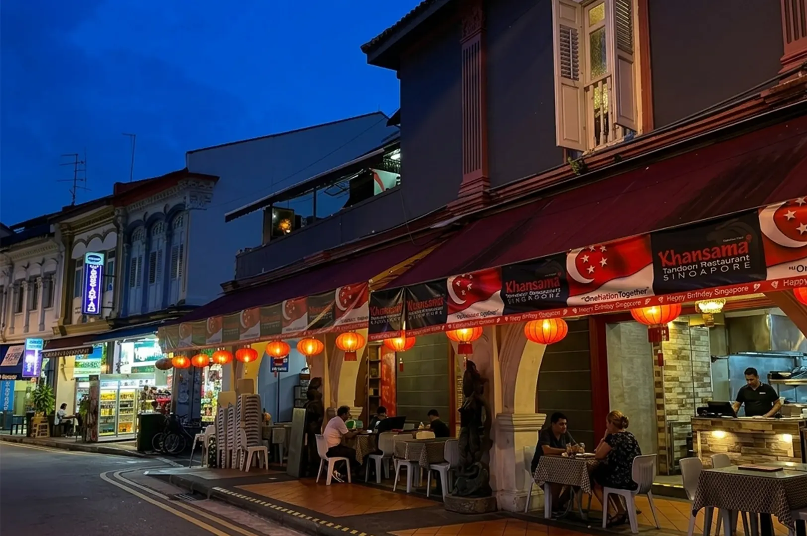 Street-side restaurant at night with red awning, lanterns, and people dining outside.