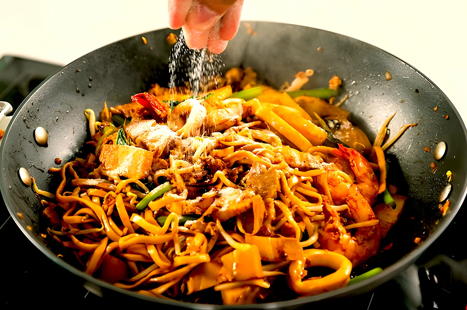 A hand sprinkles salt over sizzling noodles with shrimp and vegetables in a pan, creating a savory, appetizing dish filled with vibrant colors.