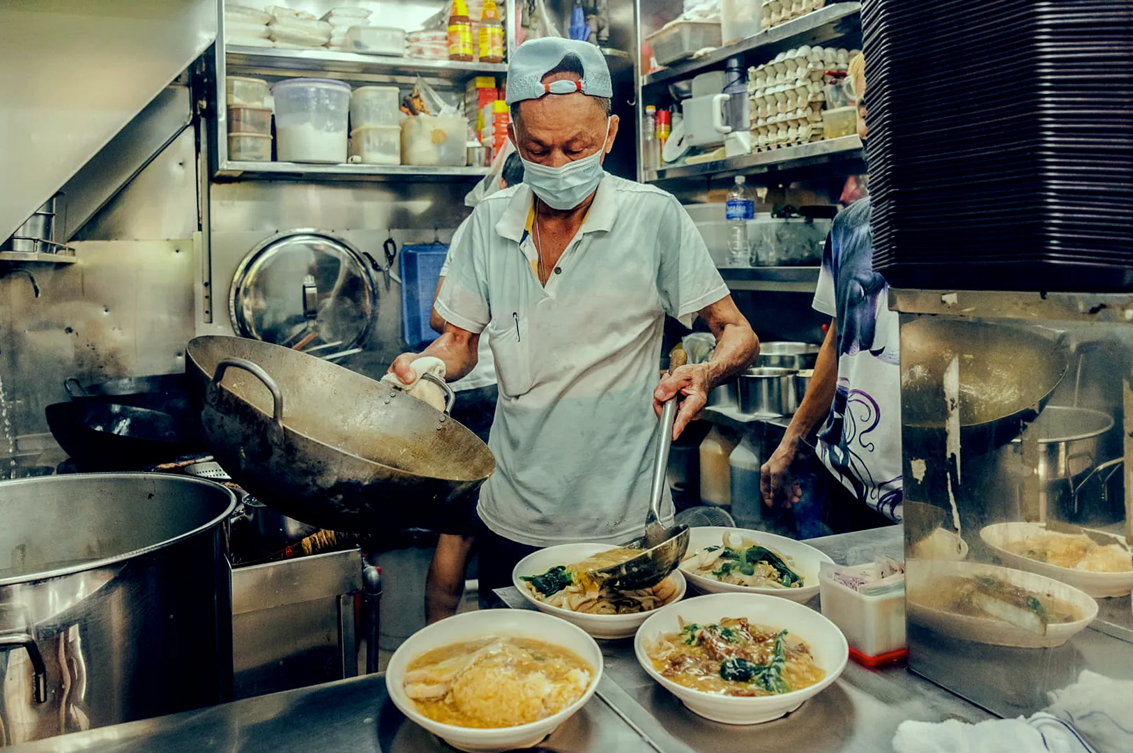 A cook in a bustling kitchen ladles soup into bowls of noodle dishes. Wearing a mask and cap, he works diligently. The kitchen is busy and full.