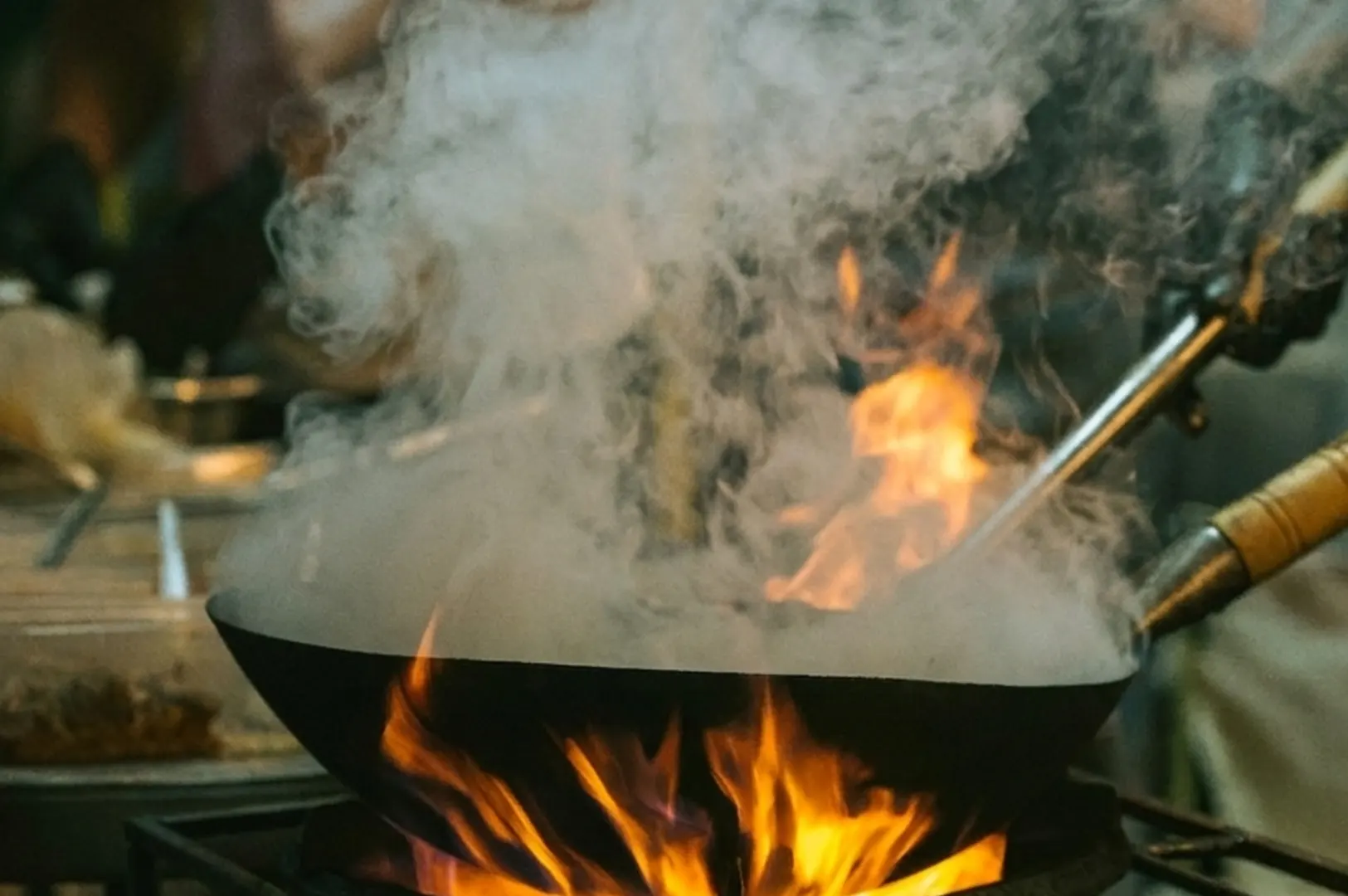 Chef tossing a wok with a stainless steel spatula over flames and smoke.