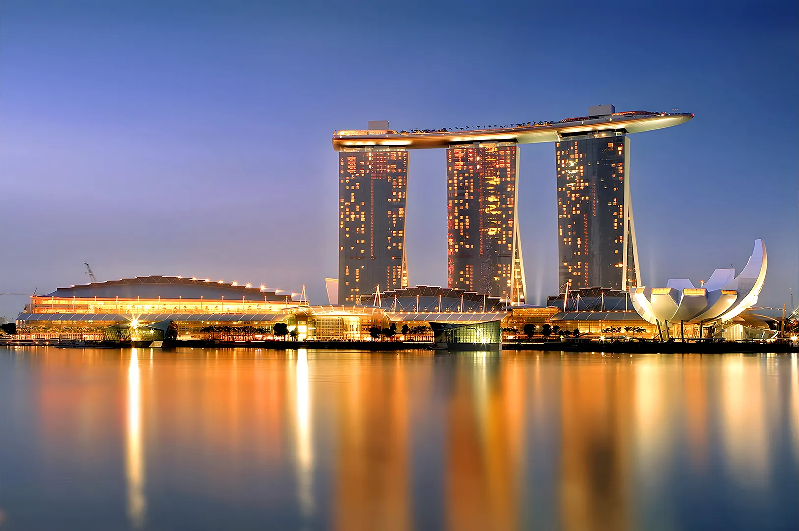 Nighttime view of Singapore's Marina Bay Sands with its three towers and connecting sky park, reflecting golden lights on the calm water.