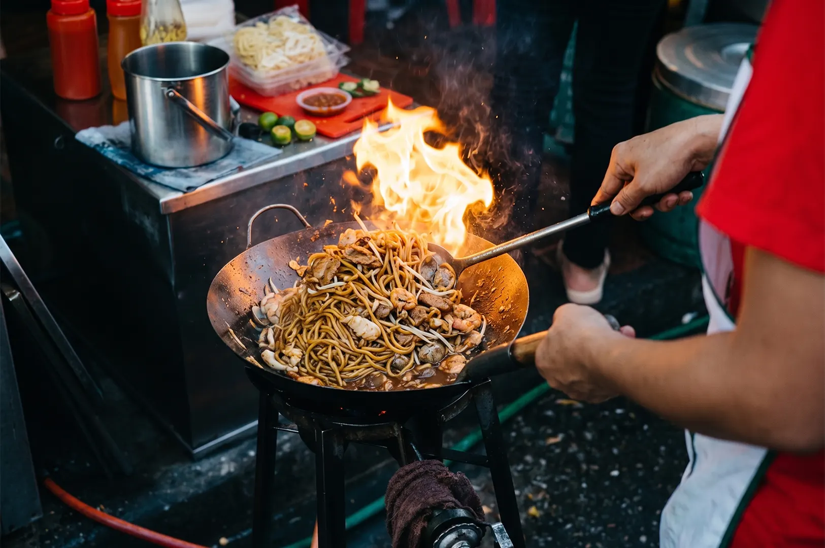 A street food chef tosses a flaming wok of stir-fried noodles with shrimp and vegetables at a night market.