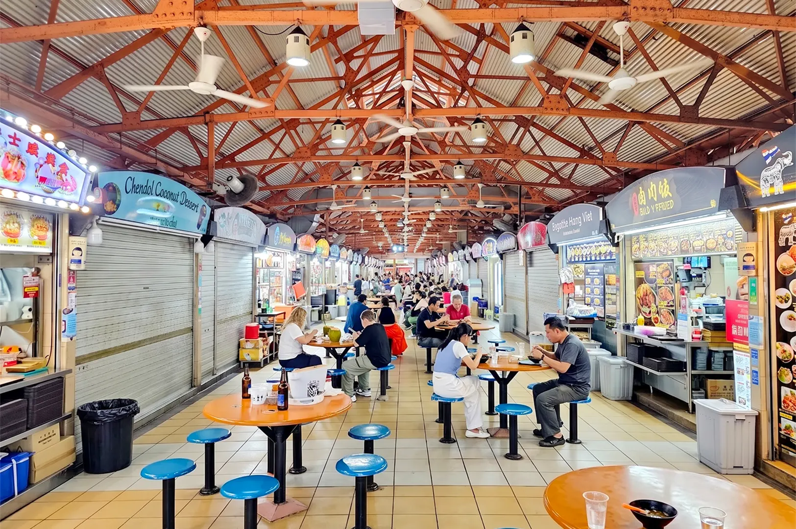 A traditional indoor hawker center or food court with various street food stalls, orange communal tables, and industrial ceiling fans in a bustling atmosphere.