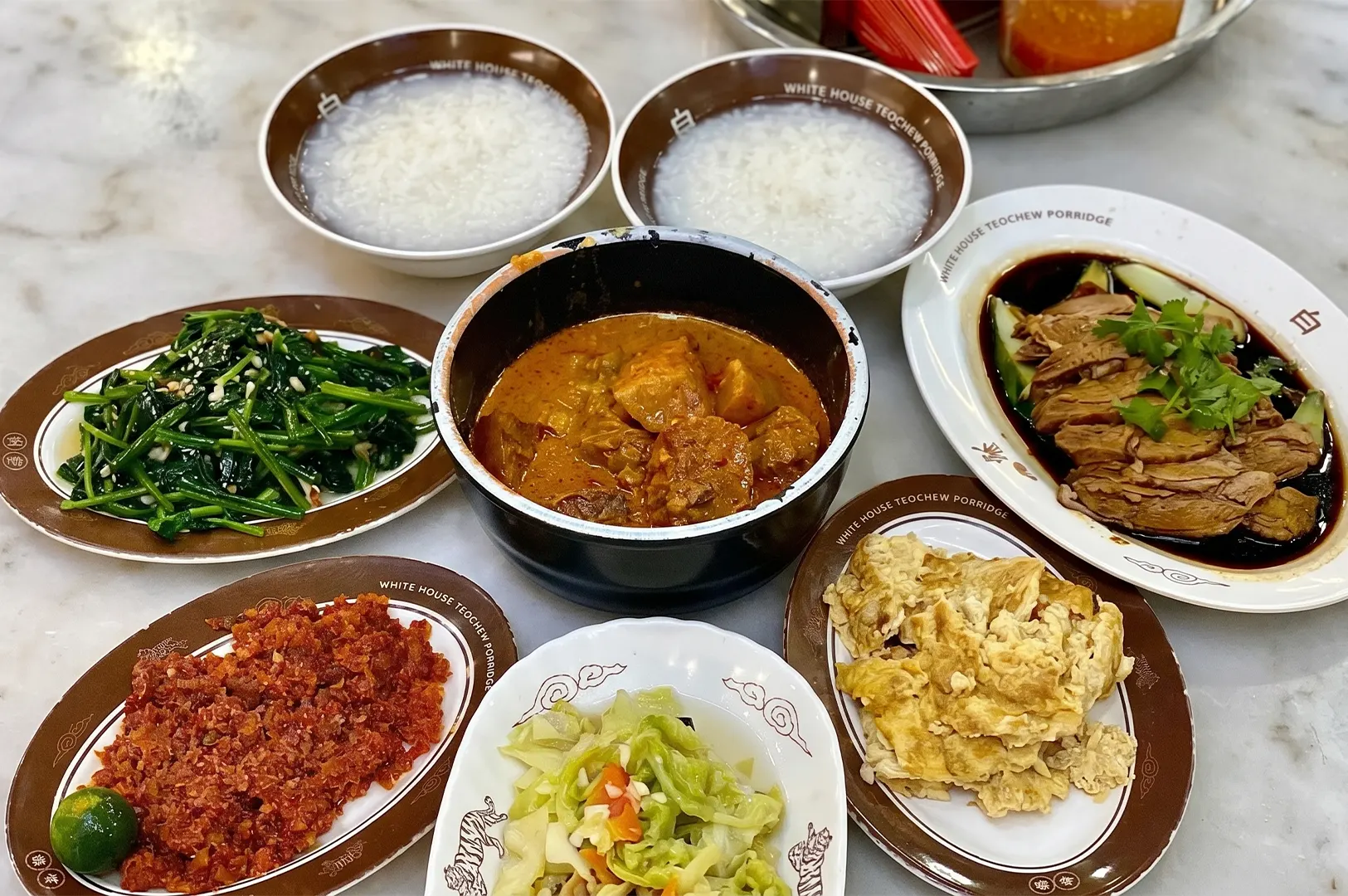 A variety of traditional Teochew porridge side dishes including braised duck, curry chicken, minced meat with chilli, stir-fried vegetables, and omelette, served with two bowls of plain rice porridge at White House Teochew Porridge.