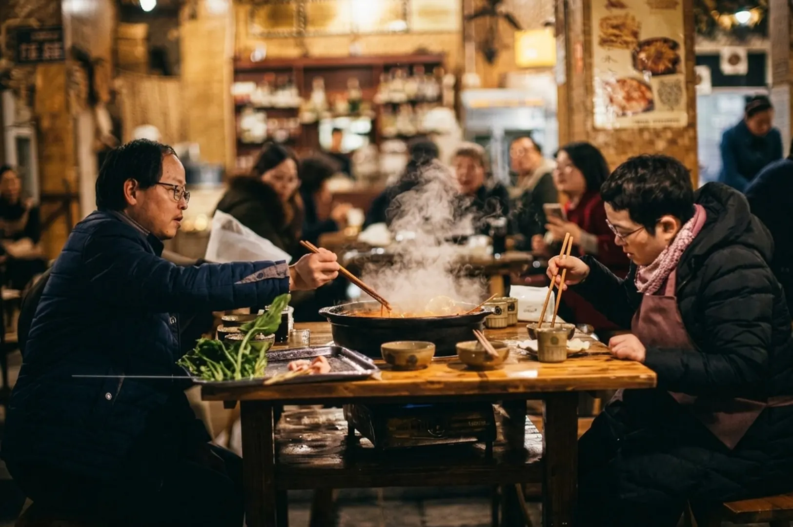 Two people eating a steaming hot pot meal with chopsticks in a cozy, crowded restaurant.