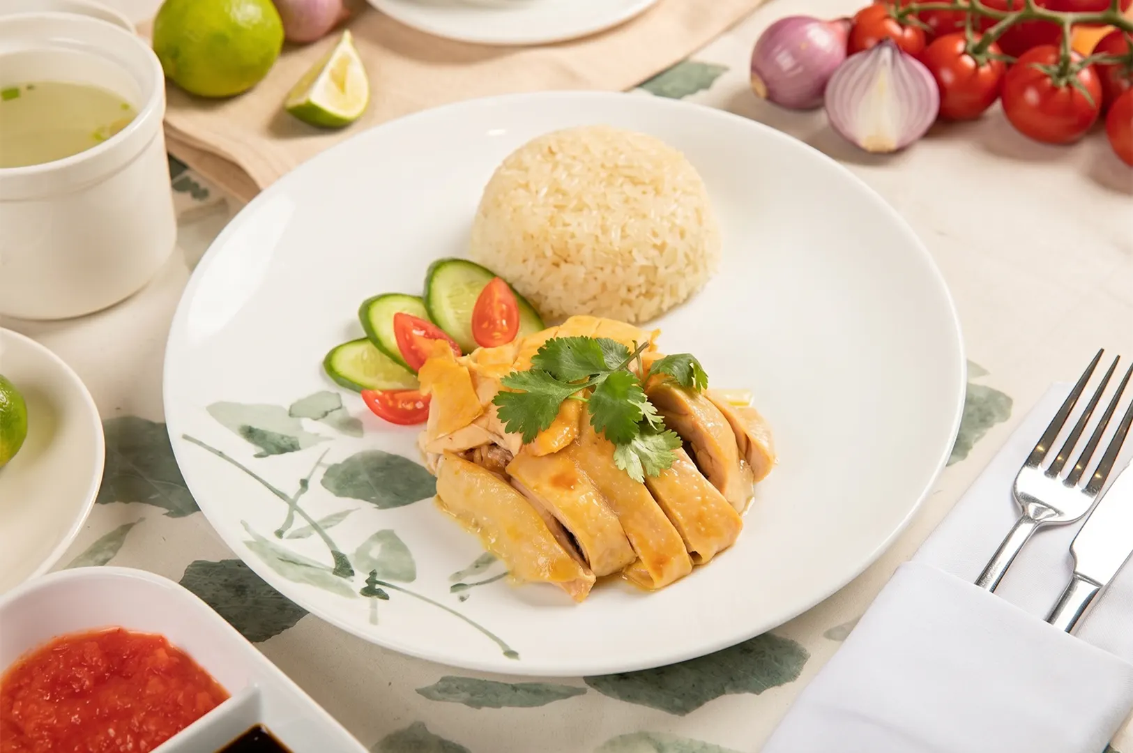 Hainanese chicken rice served with sliced cucumber, tomatoes, and a side of chili sauce.