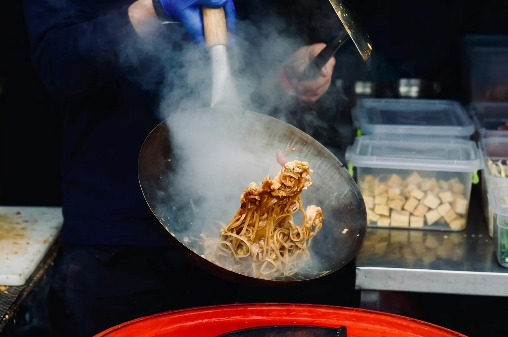 Chef tossing steaming Pad Thai noodles in a wok at a street food stall.