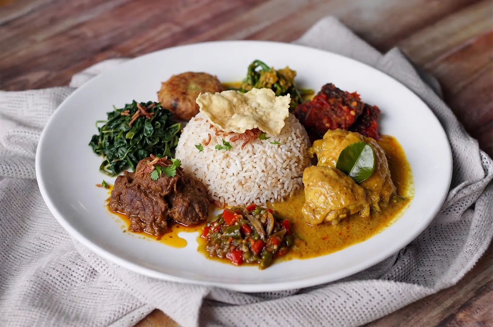 Traditional Indonesian Nasi Padang platter with beef rendang, chicken curry, sambal, and cassava leaves on a white plate.