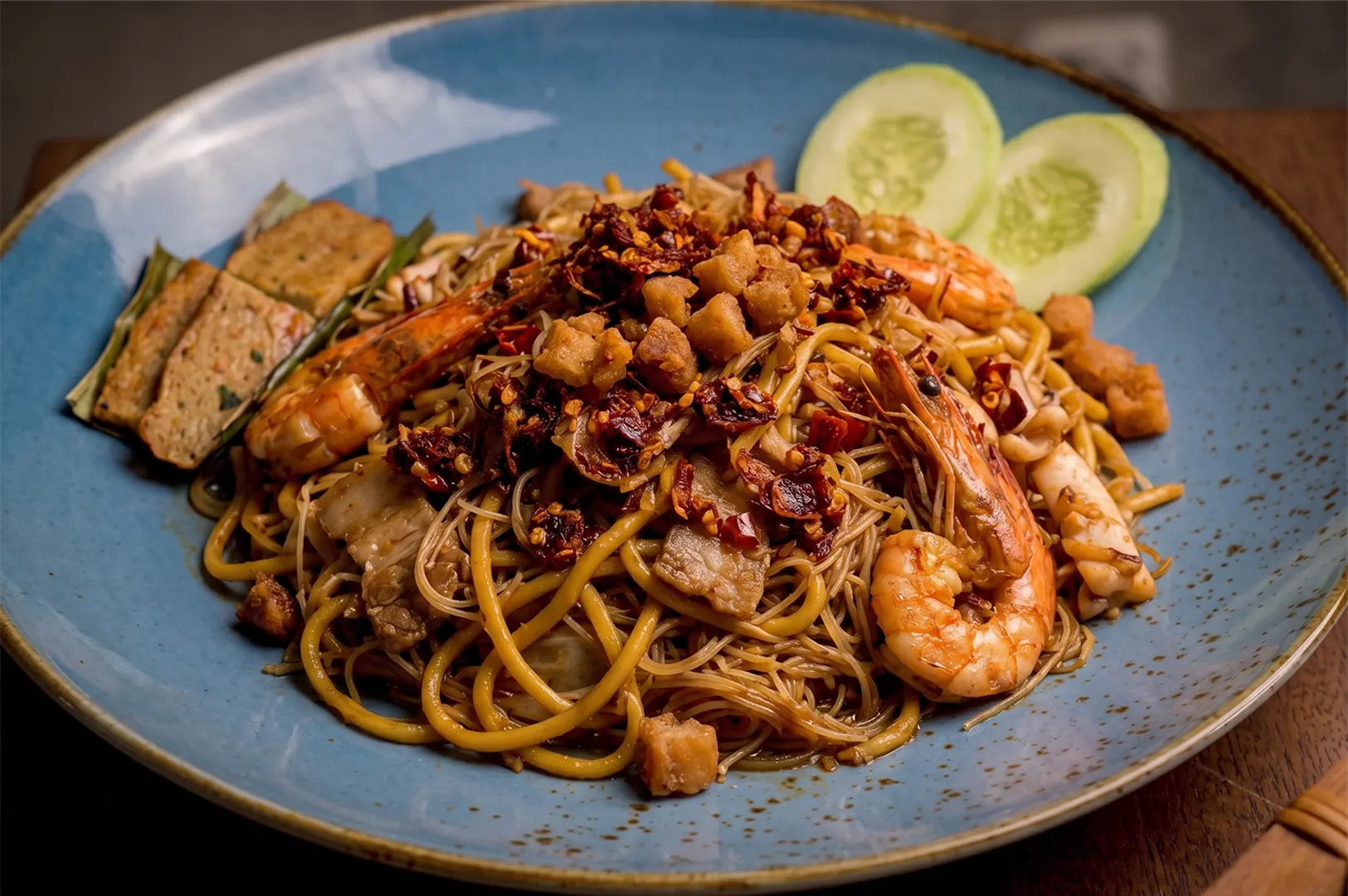 Gourmet plate of Hokkien Mee featuring thick yellow noodles, rice vermicelli, whole prawns, crispy pork lard, and chili flakes, served with cucumber slices and otah.