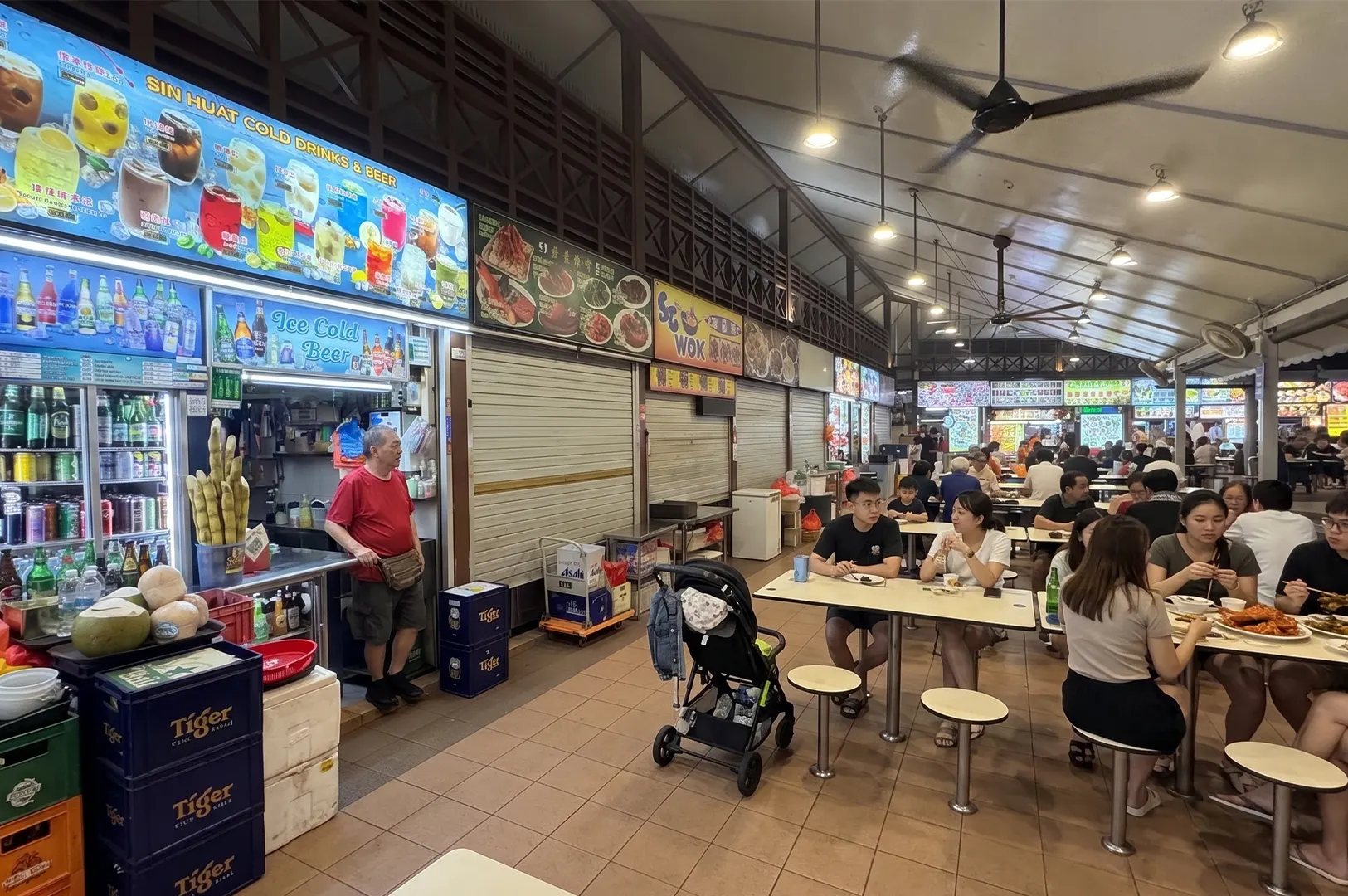 Busy outdoor hawker center in Singapore featuring a drink stall, seating area with diners, and various food kiosks under a high-ceilinged roof.