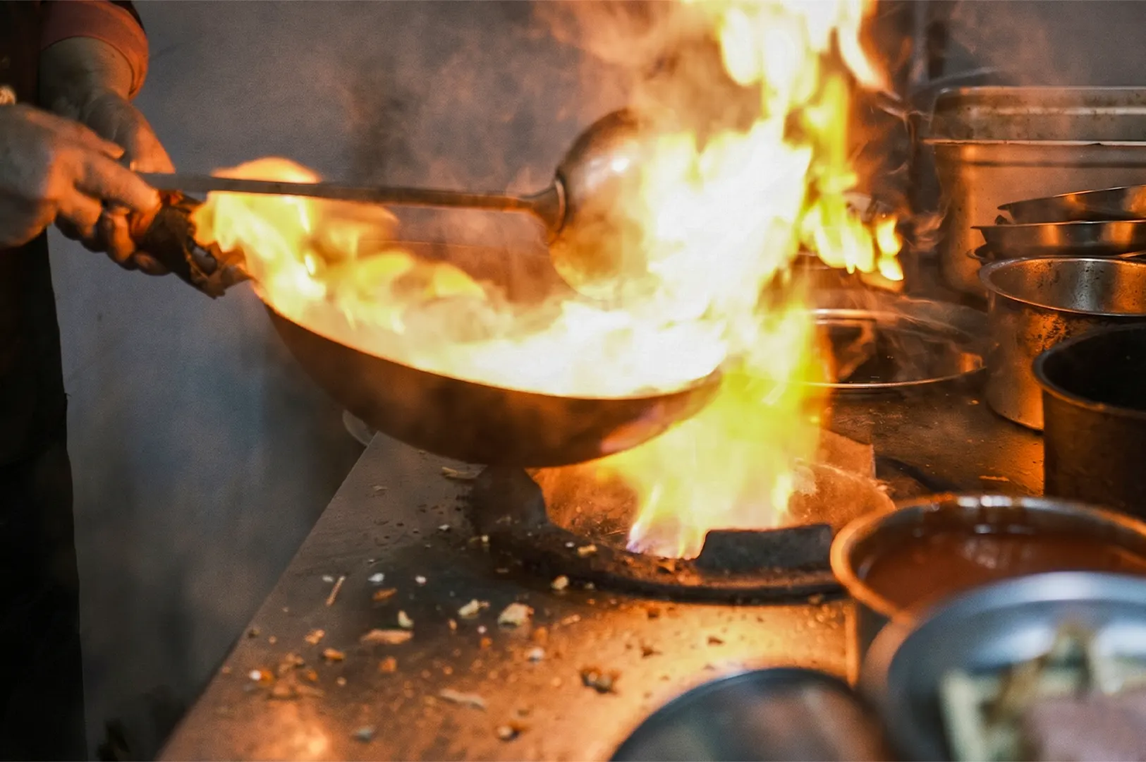 Chef stir-frying food in a wok over a high flame burner in a commercial kitchen.
