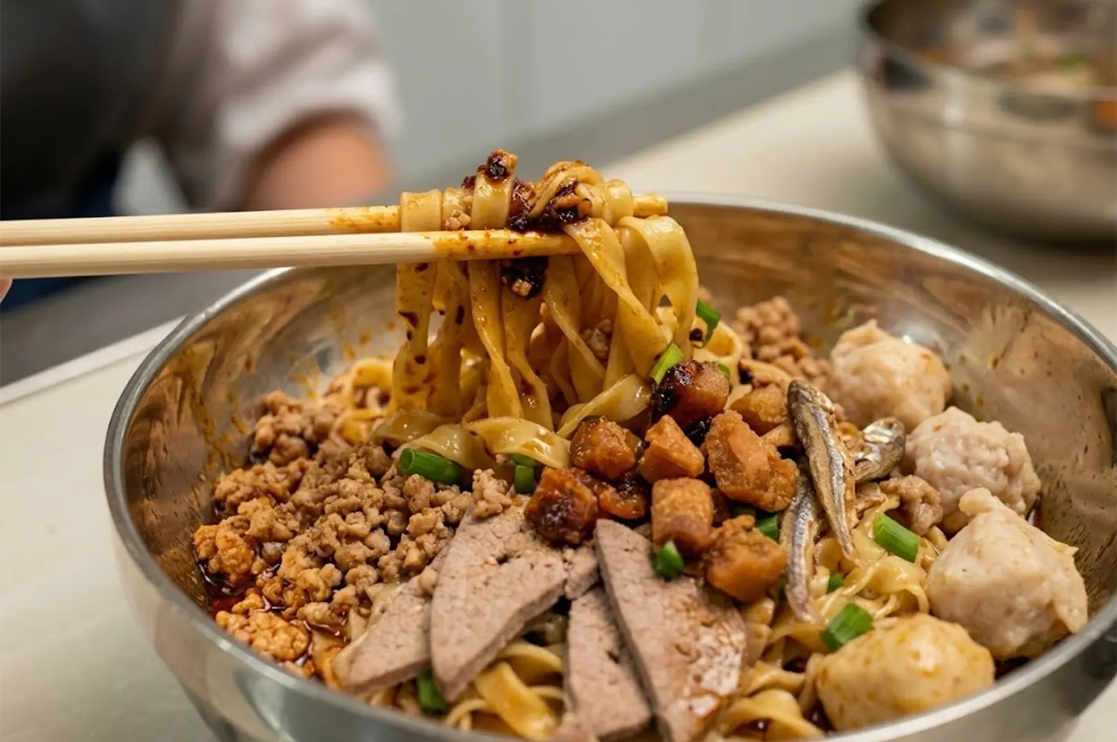 A bowl of dry Bak Chor Mee (minced meat noodles) being lifted with chopsticks, featuring flat mee pok noodles, pork liver slices, meatballs, minced pork, and crispy lard in a stainless steel bowl.