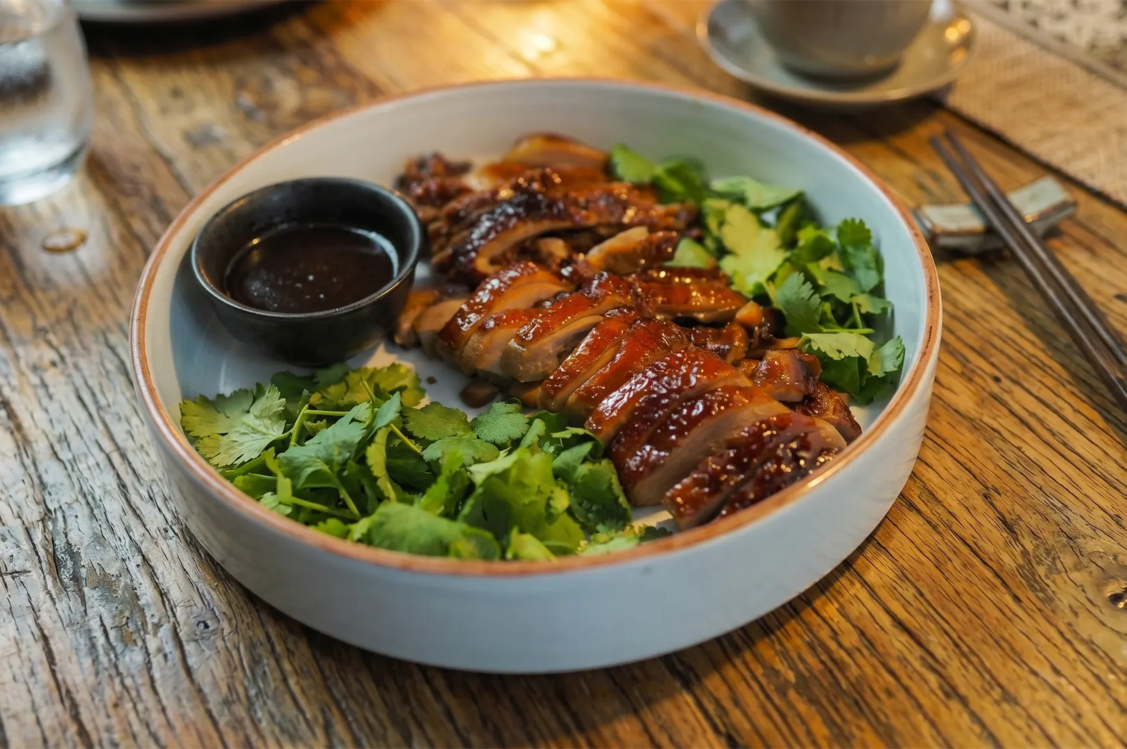 Slices of succulent roasted duck with crispy skin, garnished with fresh cilantro and served with a side of dark dipping sauce in a ceramic bowl on a rustic wooden table.