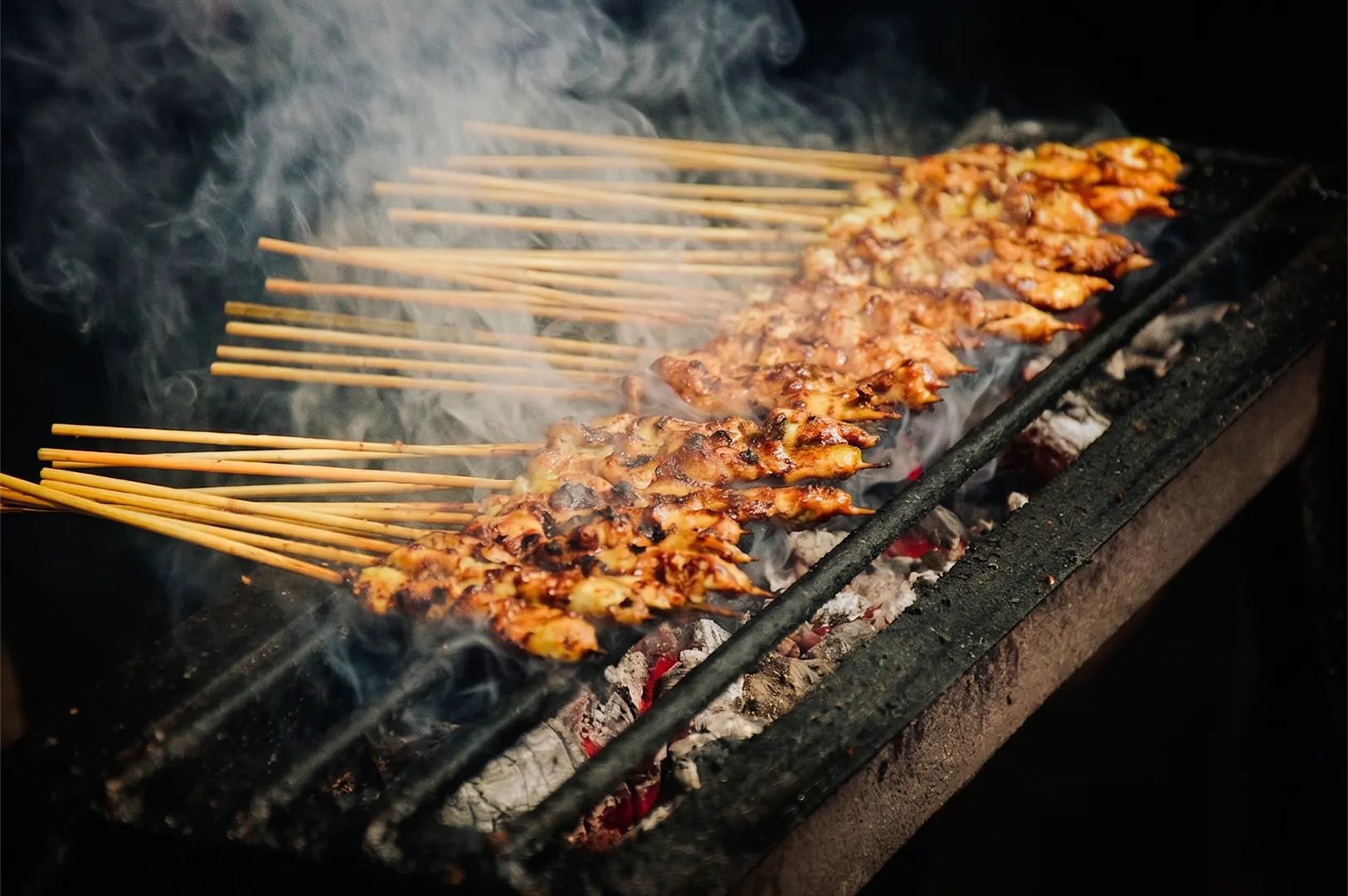 Grilled chicken satay skewers cooking over a smoky charcoal grill at a street food market.